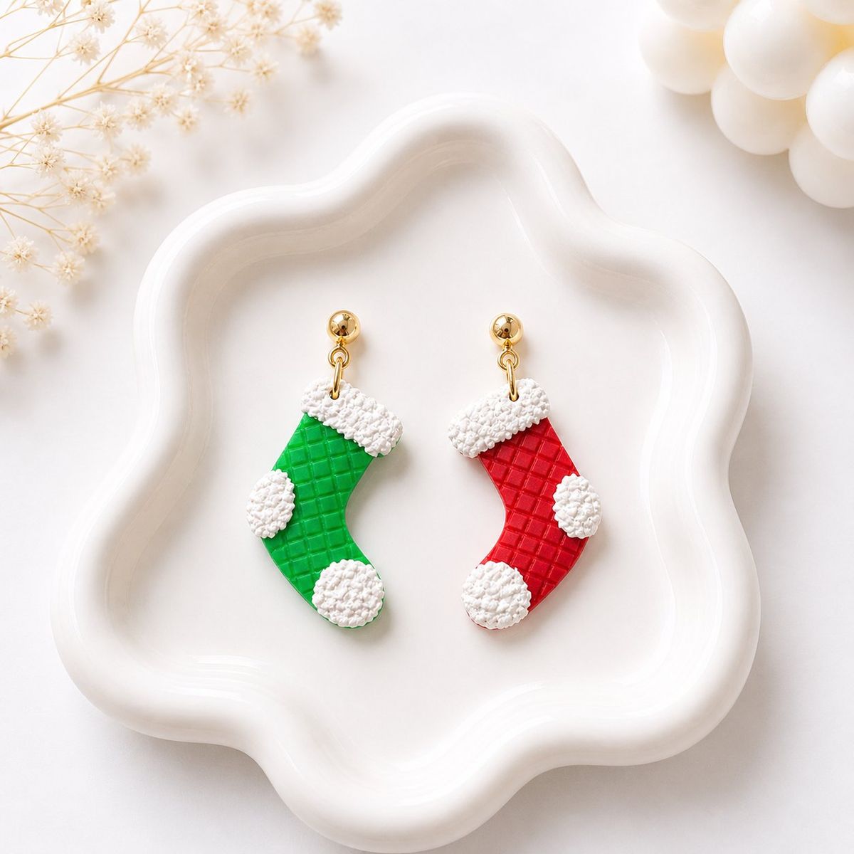 Two mismatched polymer clay Christmas stocking dangle earrings — one green and one red — with white textured trim and gold ball stud tops, displayed on a white ceramic tray.