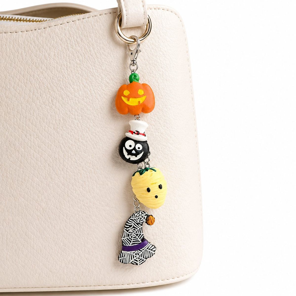 A Halloween-themed bag charm with four polymer clay charms — a jack-o'-lantern, a skull with a top hat, a mummy, and a spider web witch hat — hanging on a silver chain clipped to a cream handbag.