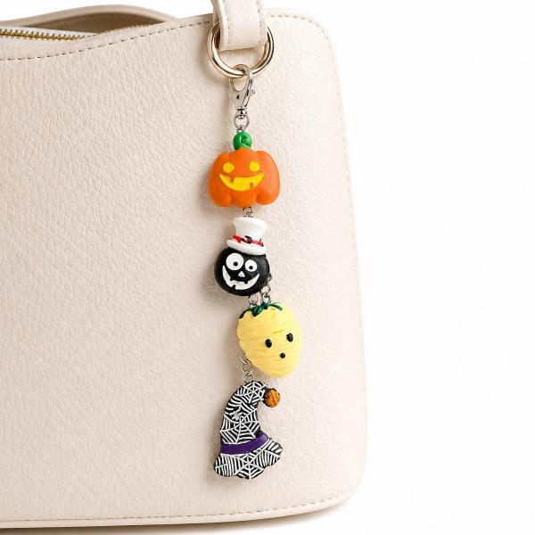 A Halloween-themed bag charm with four polymer clay charms — a jack-o'-lantern, a skull with a top hat, a mummy, and a spider web witch hat — hanging on a silver chain clipped to a cream handbag.