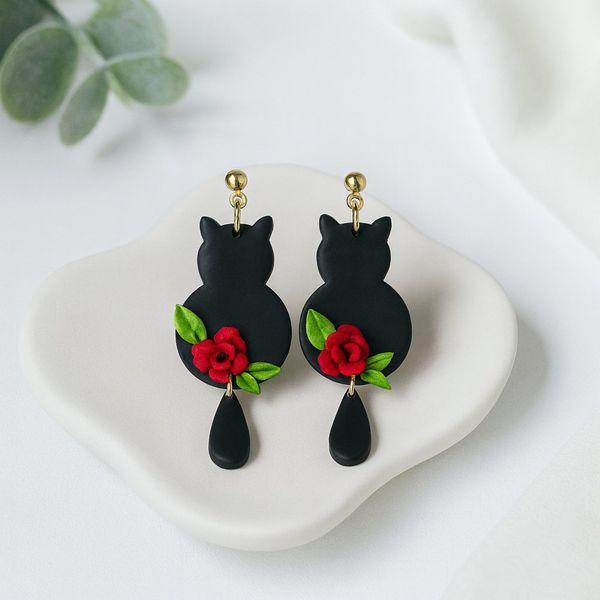 A pair of black clay cat silhouette dangle earrings with red rose and green leaf clay details and a small teardrop pendant, displayed on a white ceramic dish.