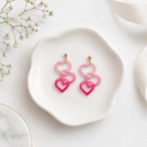 A pair of dangle earrings with two interlocked open heart charms in light pink and hot pink clay, attached to gold ball studs, displayed on a white ceramic dish.