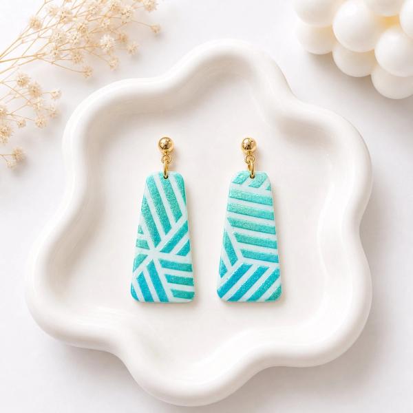 A pair of teal and white geometric patterned clay dangle earrings with gold ball studs displayed on a white wavy ceramic tray.