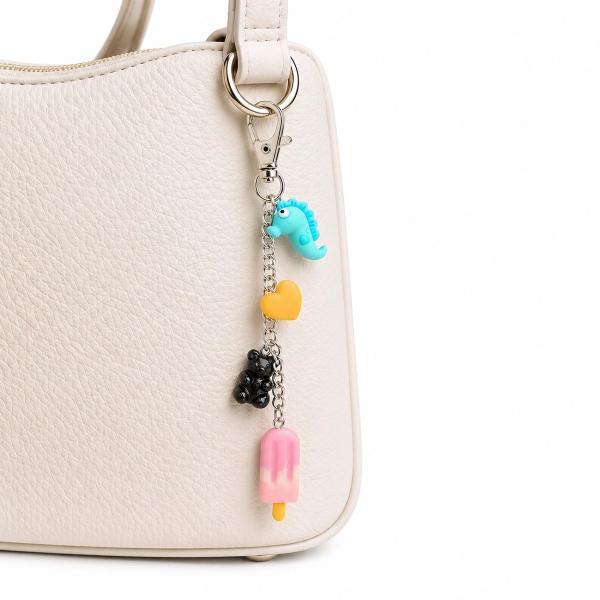 A silver lobster-clasp bag charm with four polymer clay charms — a teal dinosaur, orange heart, black gummy bear, and pink popsicle — hanging from a cream handbag.
