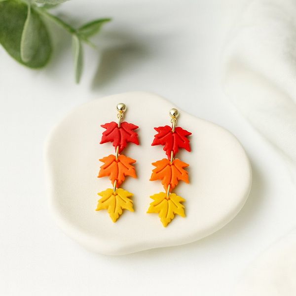 A pair of handmade clay dangle earrings with three tiered maple leaf charms in red, orange, and yellow connected by gold-tone rings, displayed on a white ceramic dish.