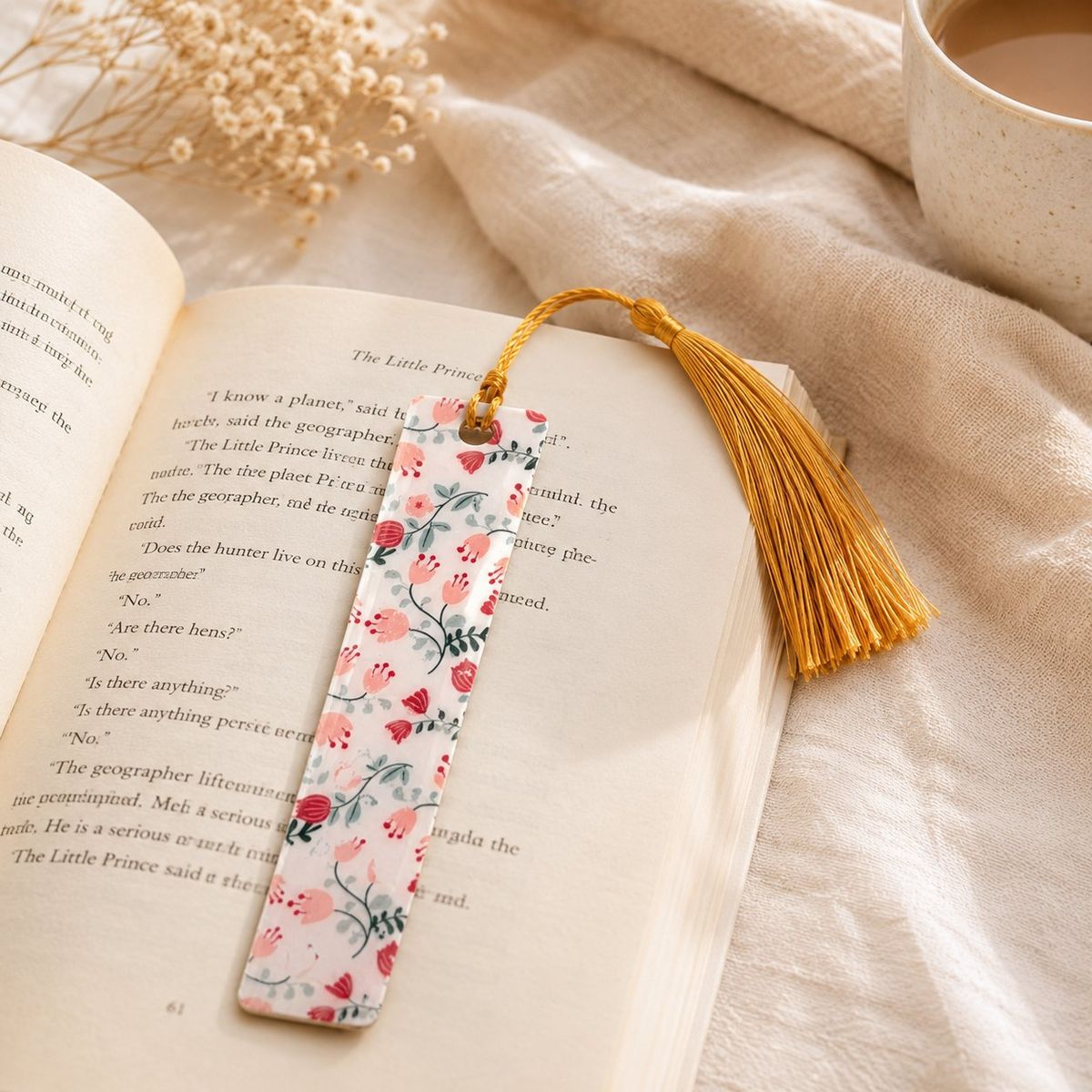 A slim white bookmark with a pink and red floral pattern and gold tassel, resting open on a copy of The Little Prince.