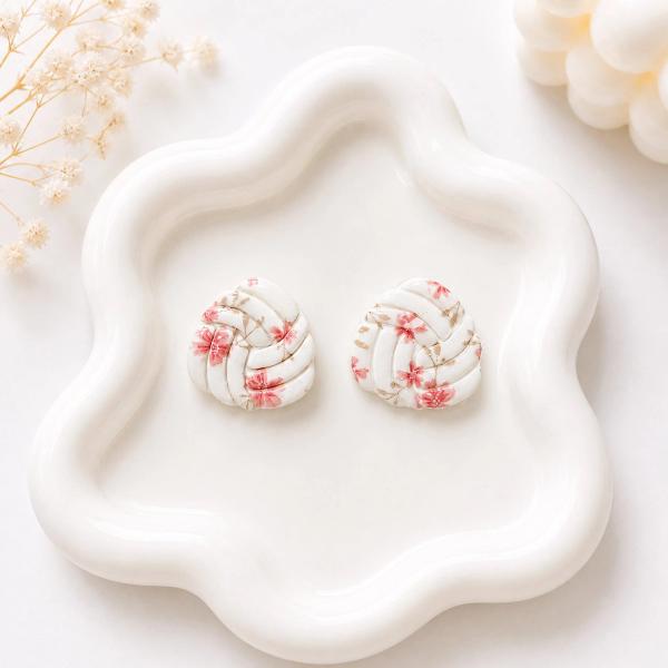 A pair of white clay knot-shaped stud earrings with a pink floral cherry blossom print, displayed on a white ceramic tray.