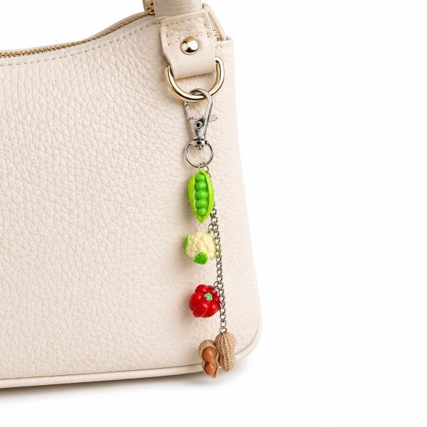 A key charm with four miniature polymer clay vegetable charms — a green pea pod, white cauliflower, red pepper, and brown peanut — hanging on a silver chain attached to a lobster clasp on a cream handbag.