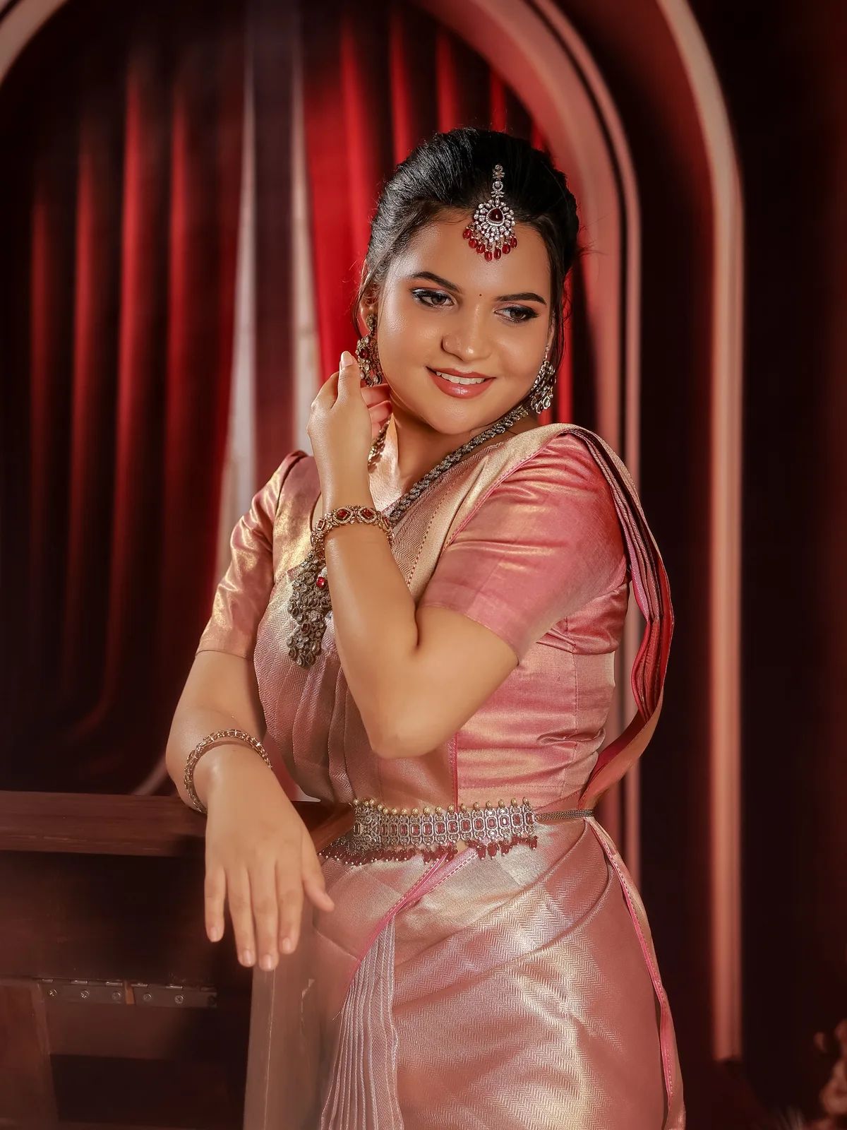 Reception bride in a rose-gold tissue saree against a crimson curtained arch