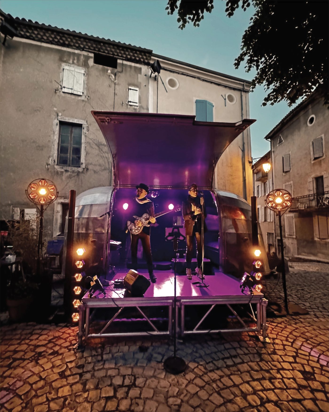 La caravane Airstream illuminée de violet, de nuit, dans une ruelle pavée de village