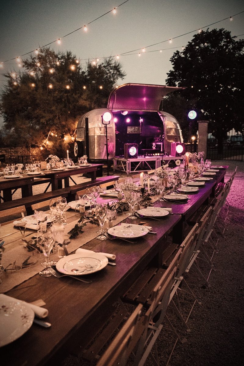 Table de réception de mariage dressée au premier plan, caravane Airstream illuminée en arrière-plan de nuit