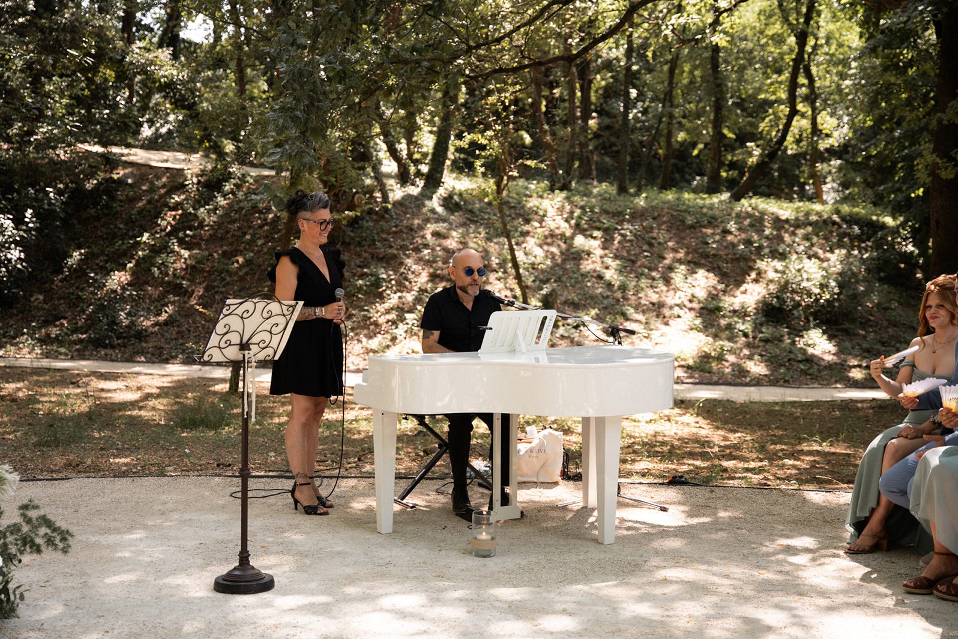 Sève, officiante, au micro à côté d'Alex jouant du piano à queue blanc lors d'une cérémonie laïque en extérieur