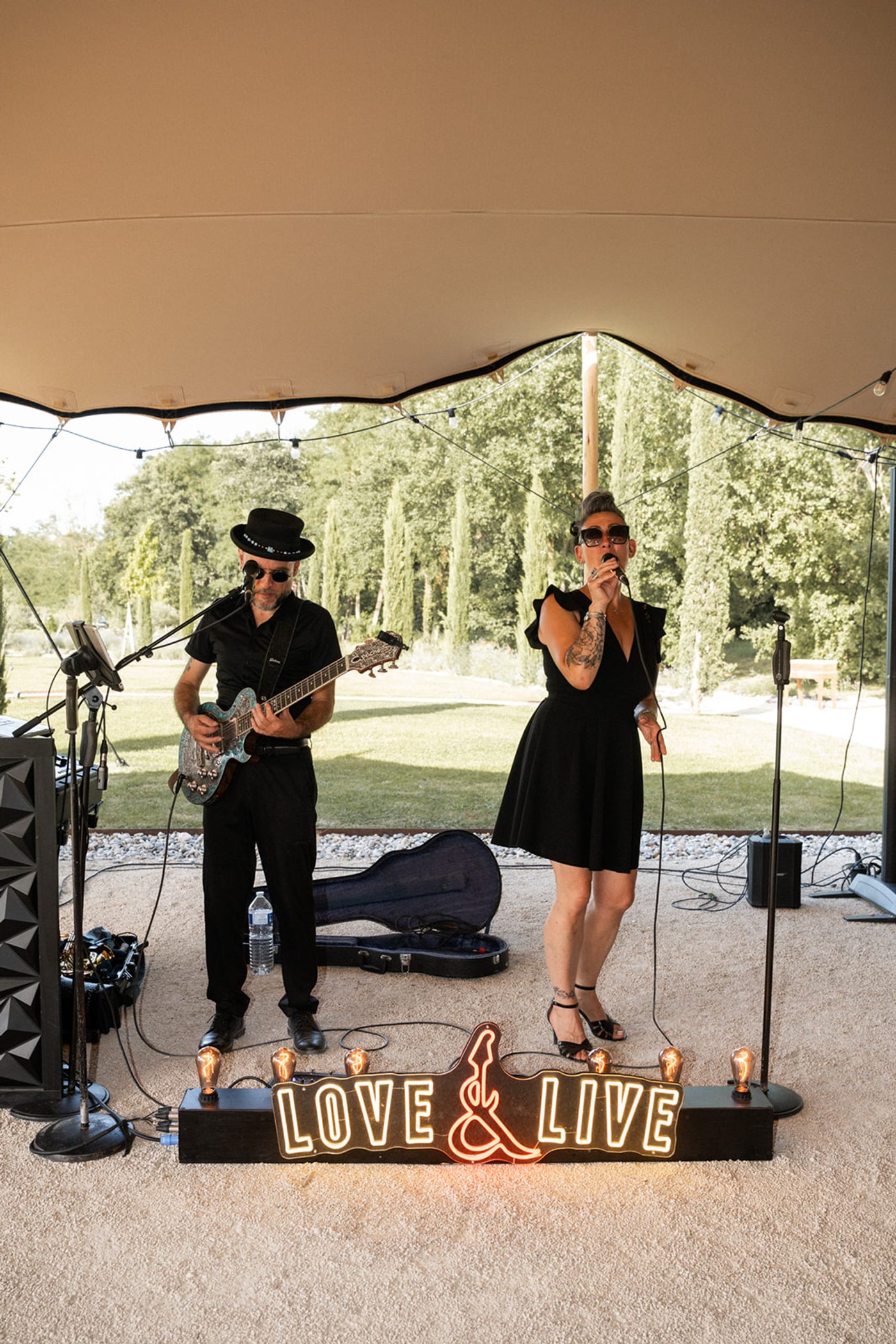 Love & Live en concert sous une voile d'ombrage, avec l'enseigne lumineuse à leurs pieds