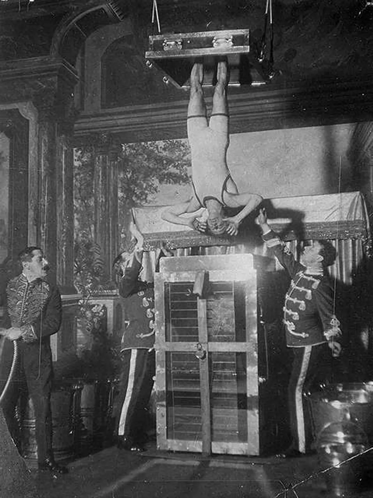 Houdini henger opp-ned foran Chinese Water Torture Cell med assistenter rundt seg.