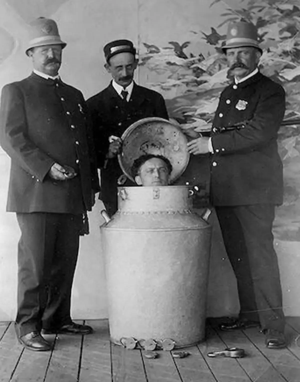 Houdini låst inne i et melkespann av metall, med hodet så vidt over kanten, flankert av to politimenn. Library of Congress.