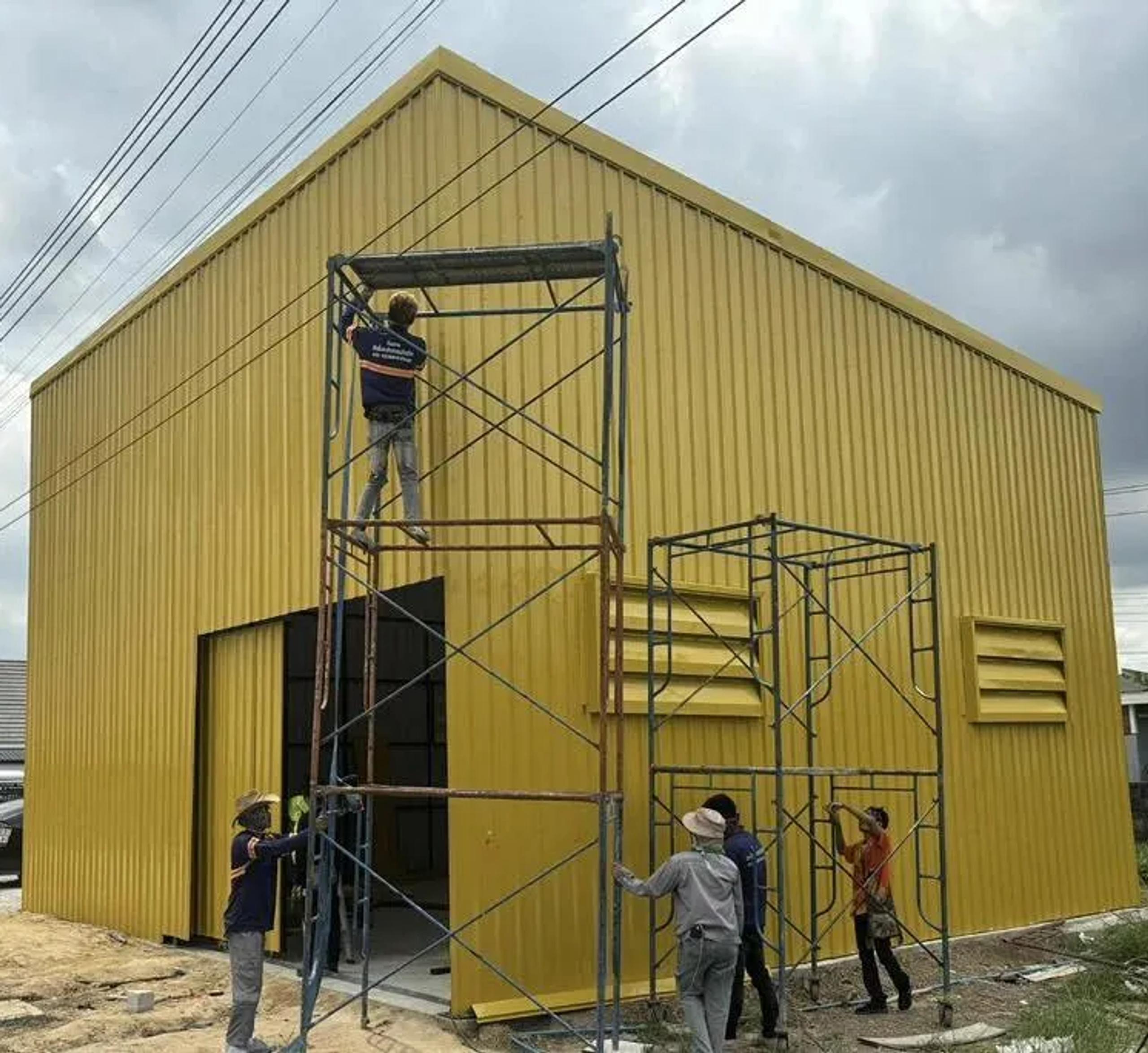 ทีมช่างติดตั้งแผ่นเหล็กสีเหลืองบนโครงสร้างโรงงานระหว่างการก่อสร้าง | Installers fitting yellow steel cladding onto a factory structure under construction