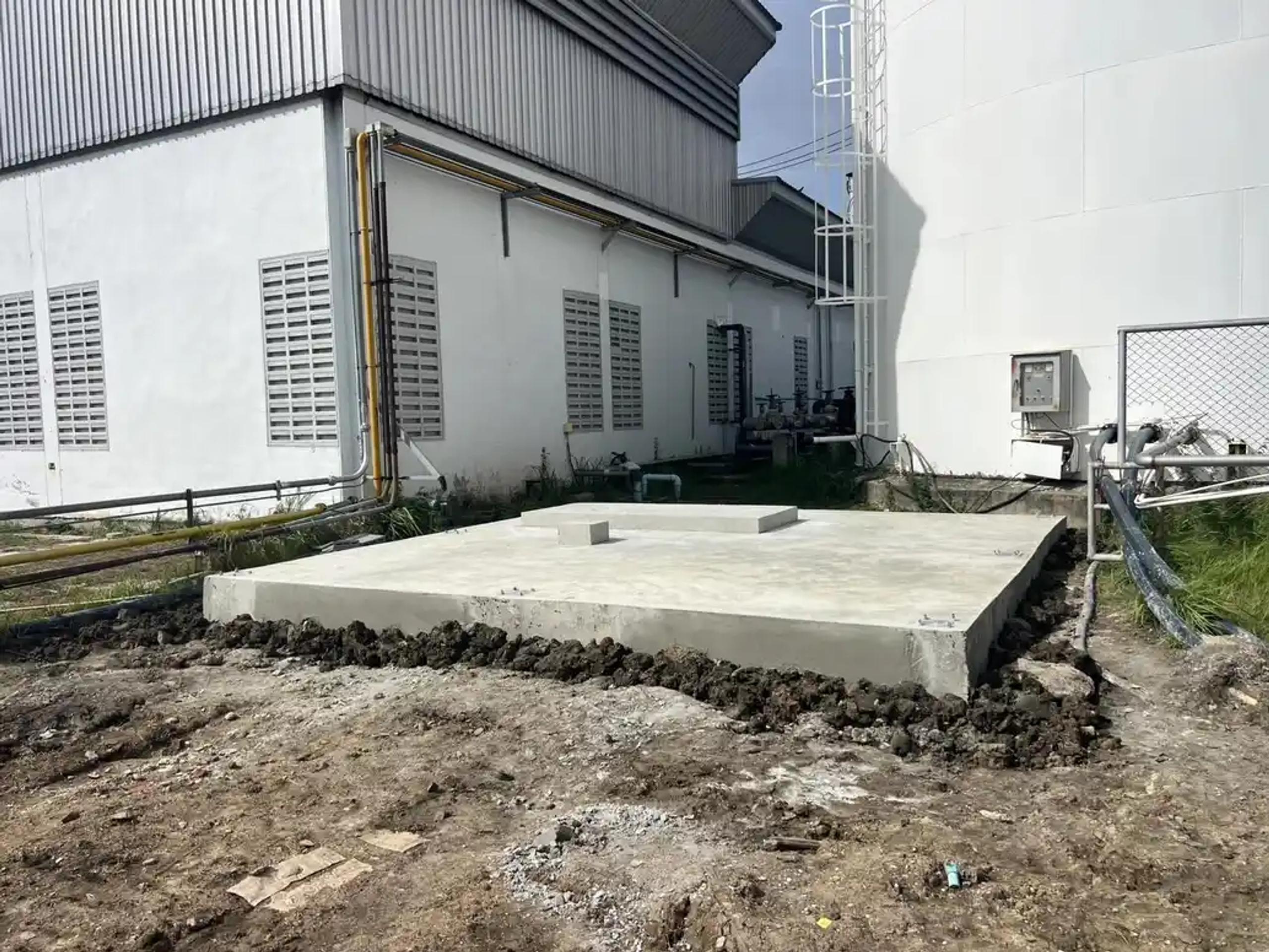 ฐานคอนกรีตที่เทเสร็จข้างอาคารโรงงานและถังเก็บสาร | Finished concrete foundation beside a factory building and storage tanks