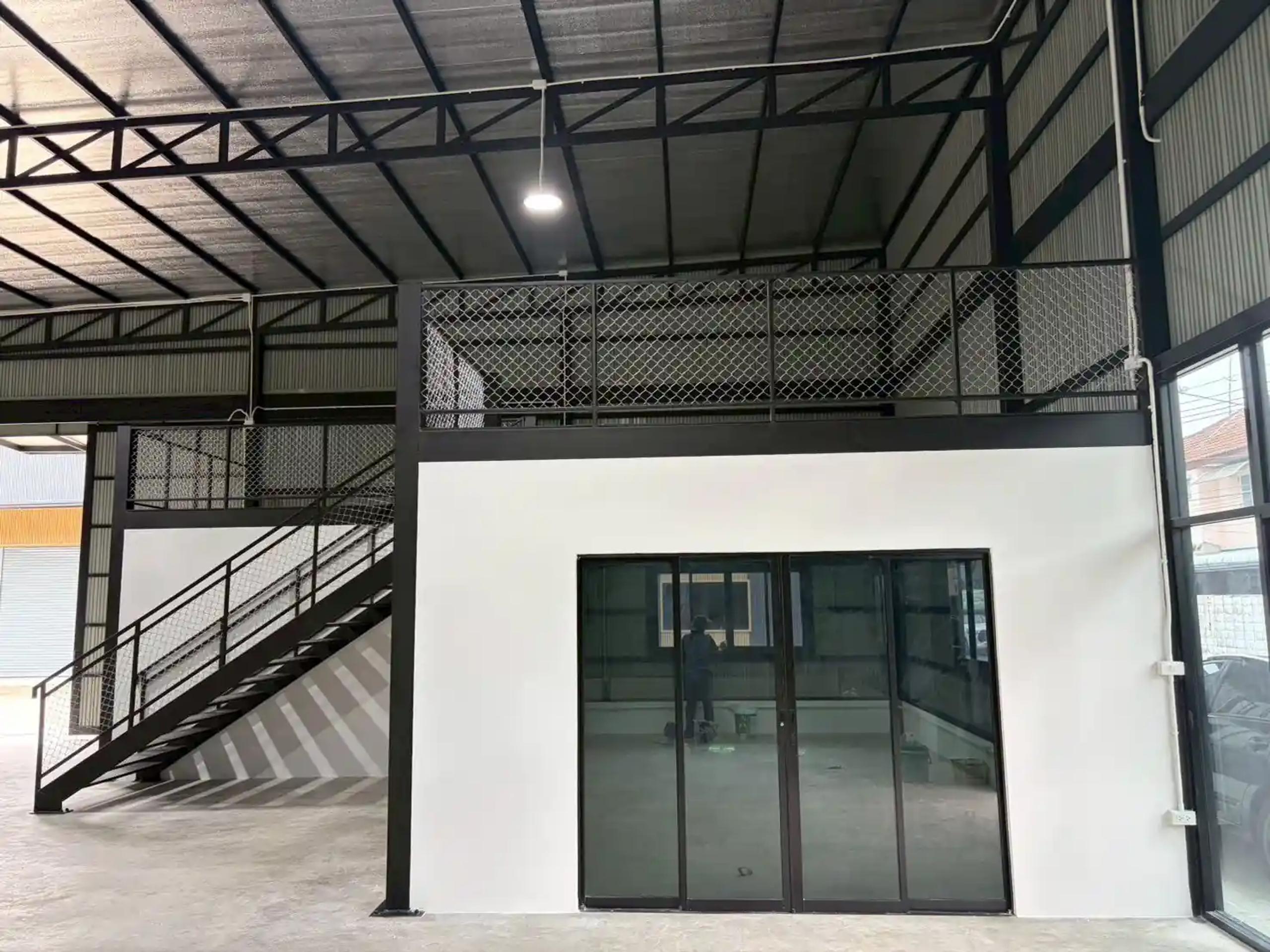 ห้องสำนักงานผนังขาวใต้ชั้นลอยโครงเหล็ก พร้อมประตูบานเลื่อนกระจกและบันไดเหล็กขึ้นชั้นลอย | White-walled office room beneath a steel mezzanine, with a sliding glass door and a steel stair to the upper level