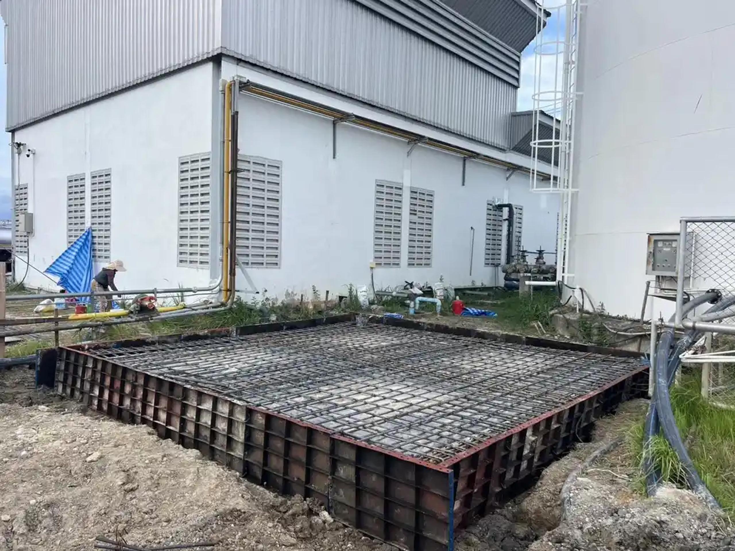 แบบหล่อและเหล็กเสริมพื้นฐานรากก่อนเทคอนกรีตข้างอาคารโรงงาน | Steel formwork and rebar for a foundation slab before pouring, beside a factory building