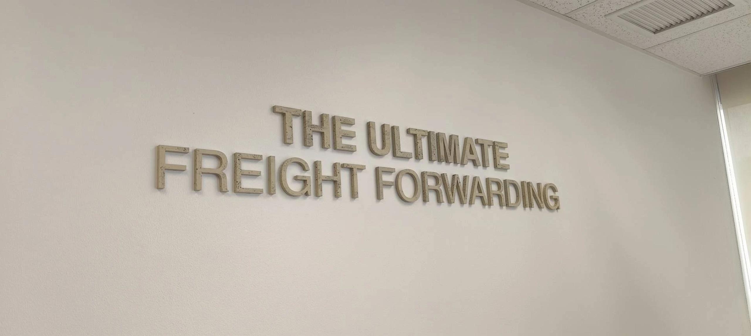 ป้ายตัวอักษรโลหะไม่มีไฟข้อความ THE ULTIMATE FREIGHT FORWARDING ติดผนังภายในสำนักงาน | Unlit dimensional letter sign reading THE ULTIMATE FREIGHT FORWARDING mounted on an office interior wall