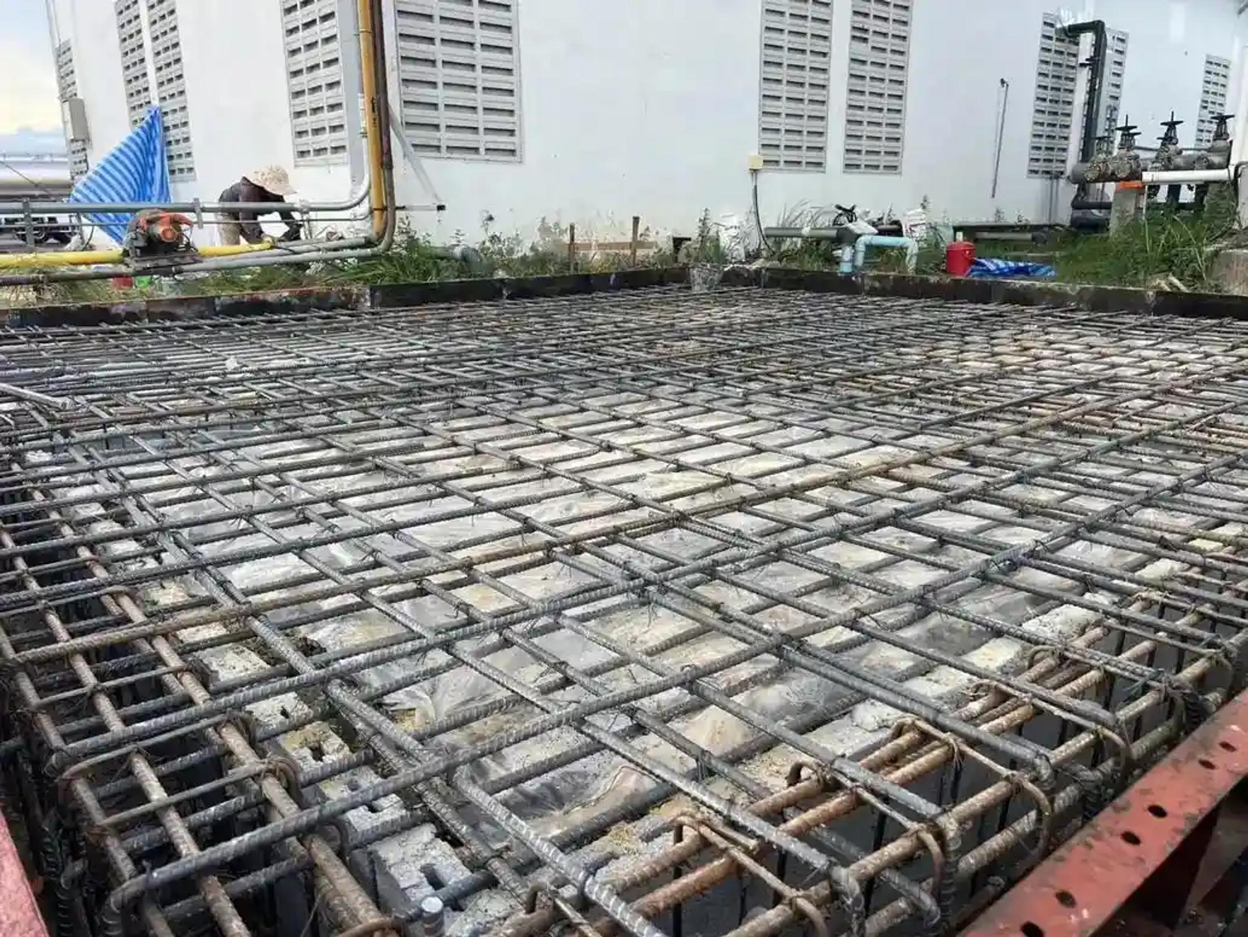 งานผูกเหล็กตะแกรงพื้นฐานรากขนาดใหญ่ก่อนเทคอนกรีตข้างอาคารโรงงาน | Large rebar mesh grid tied for a foundation slab before pouring, beside a factory building