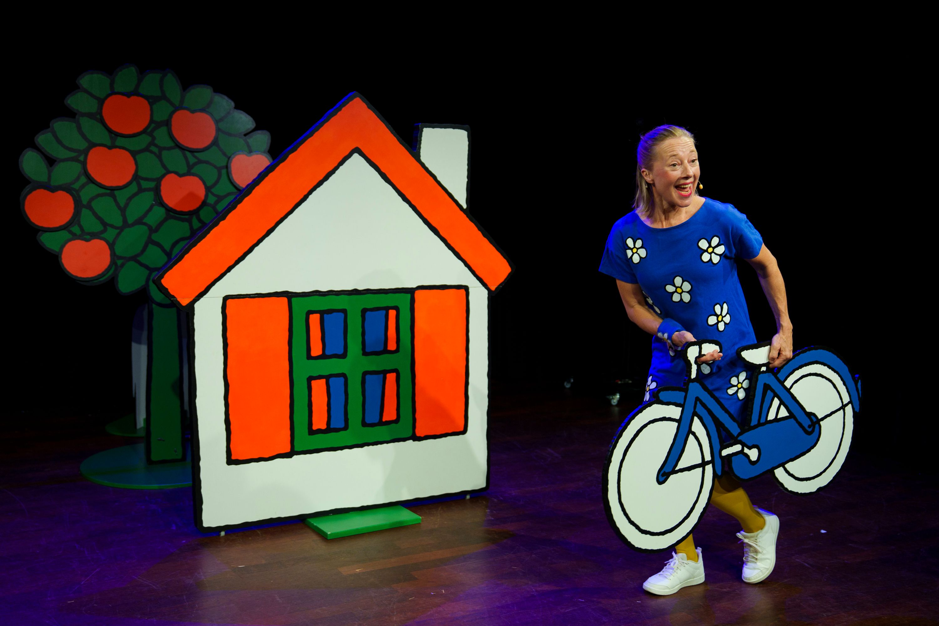 vrouw met kartonnen fiets op podium naast huis en appelboom