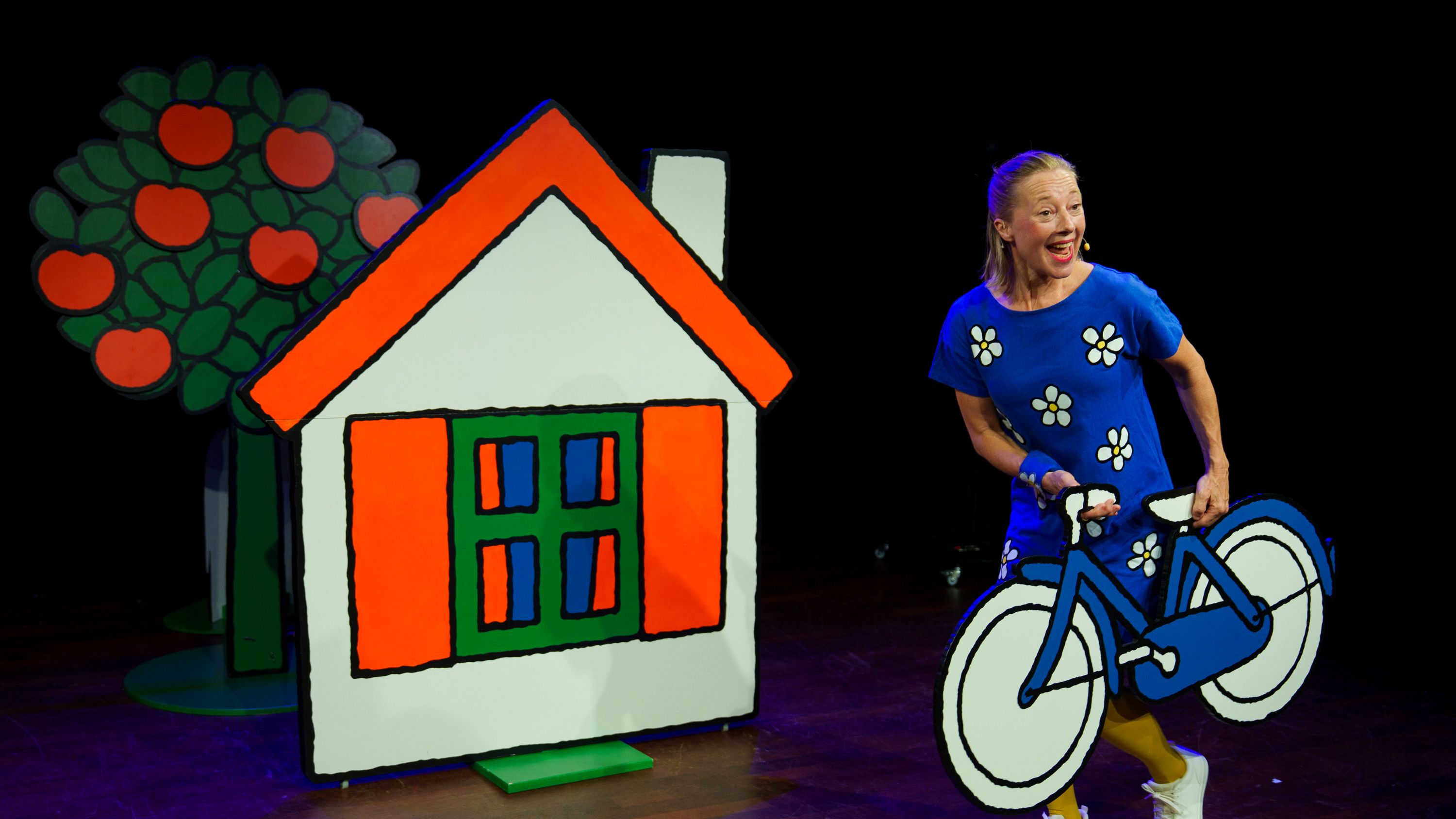 vrouw met kartonnen fiets op podium naast huis en appelboom