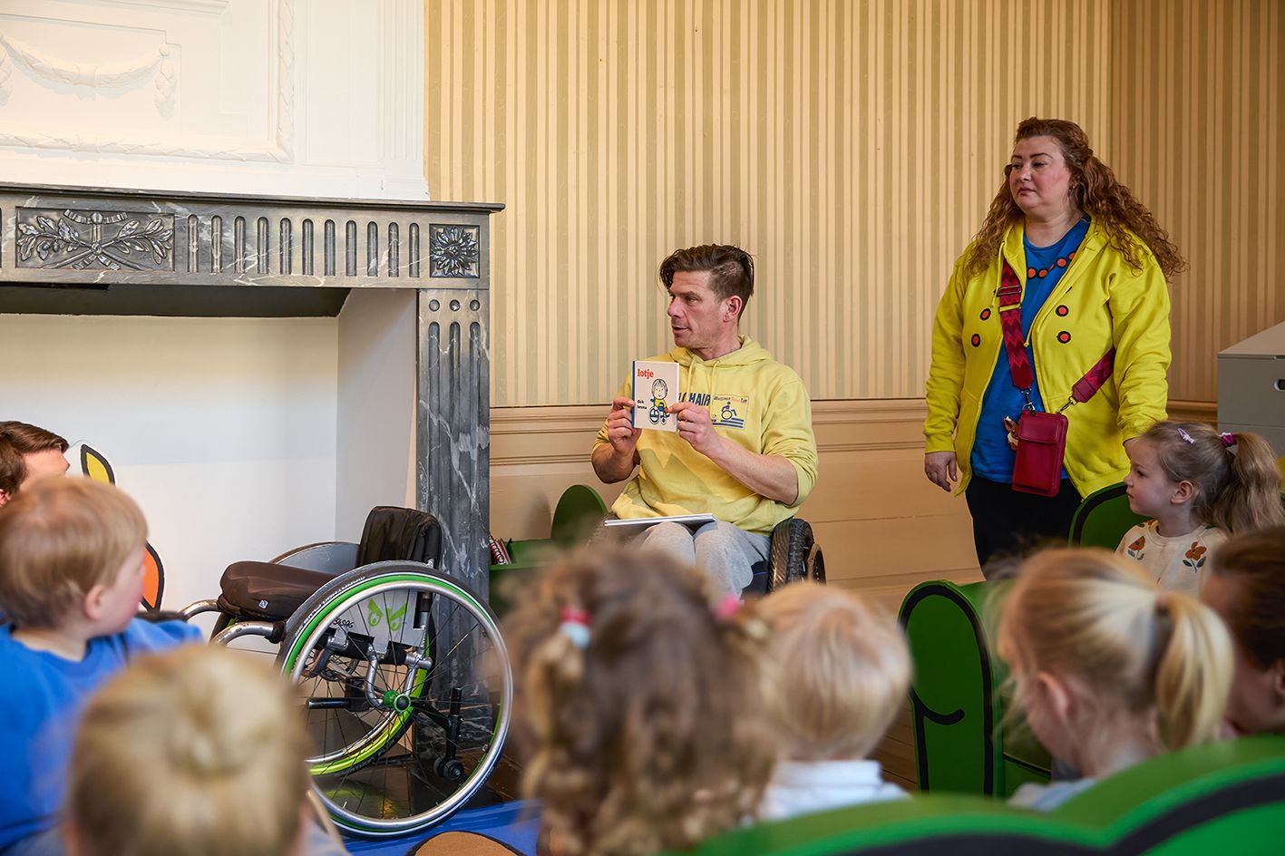 voorleessessie met kinderen en begeleiders in leeszaal. Persoon in rolstoel toont boek.