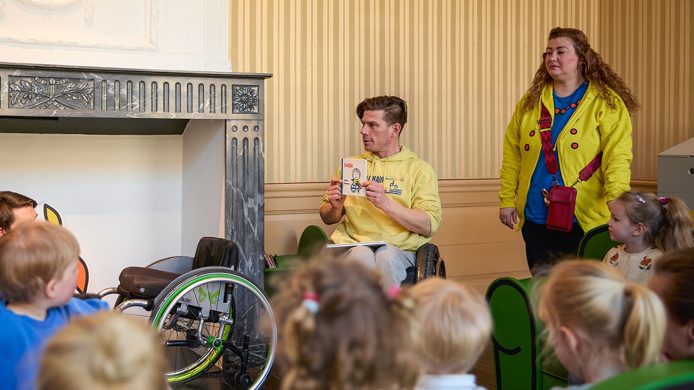 voorleessessie met kinderen en begeleiders in leeszaal. Persoon in rolstoel toont boek.