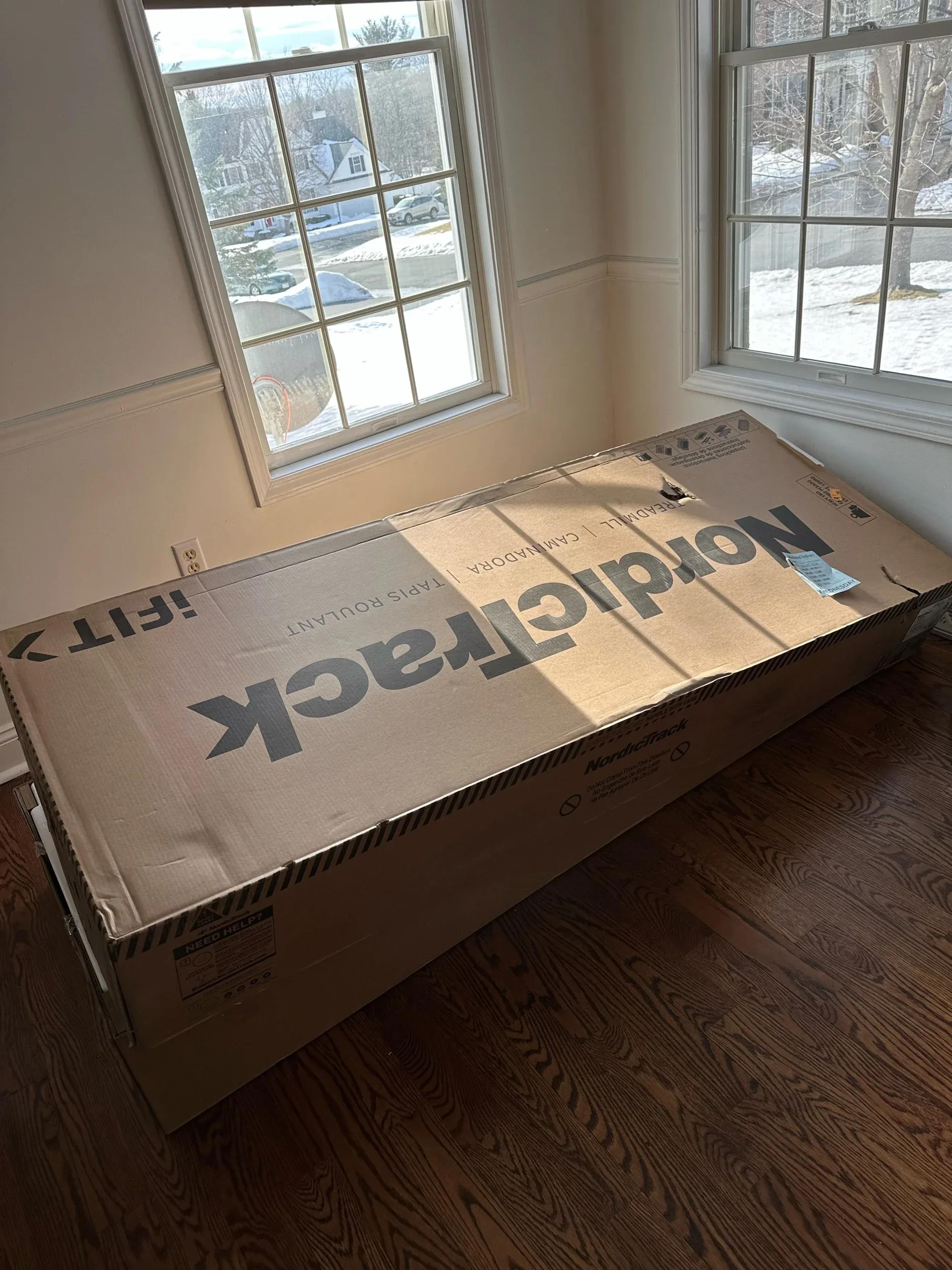 Large cardboard box labeled NordicTrack treadmill placed on hardwood floor near two windows with snow visible outside
