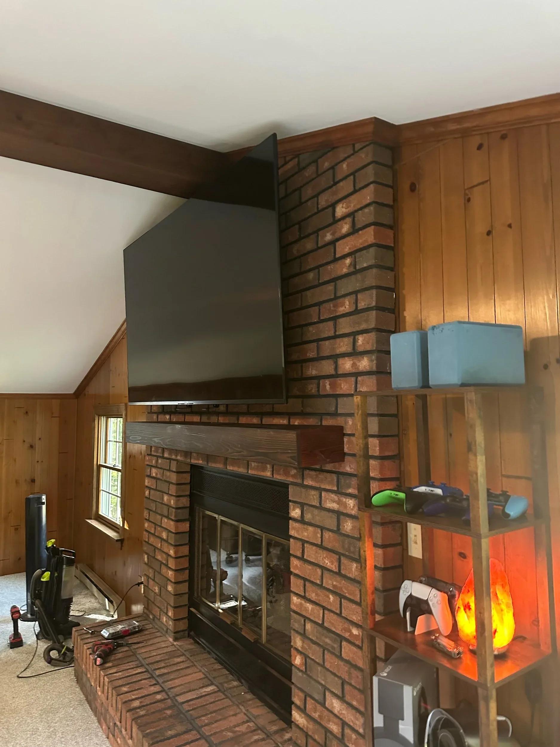 Flat-screen TV mounted on a brick chimney corner above a wooden mantel with gaming controllers on a nearby shelf