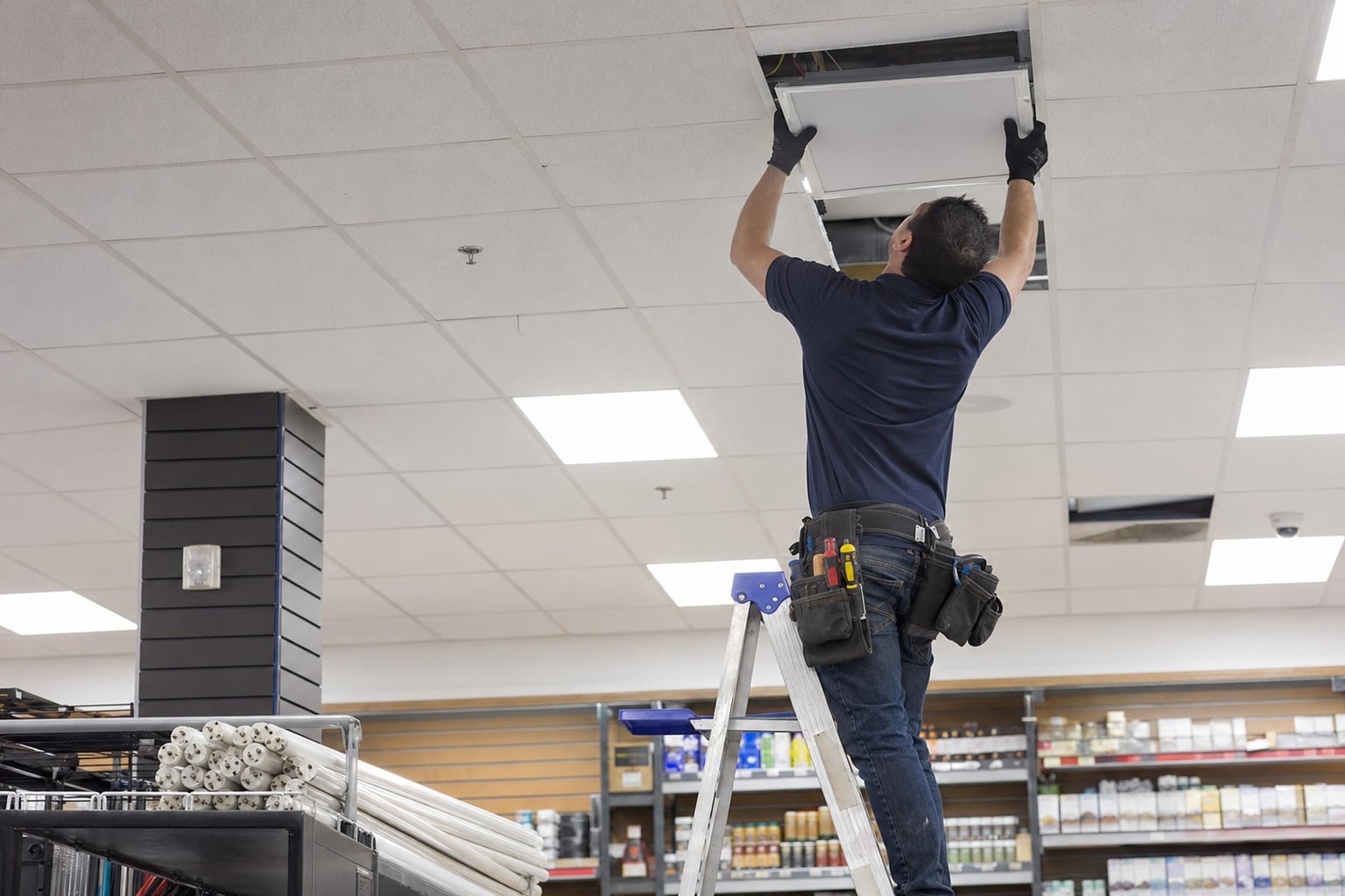 Electrician on a stepladder replacing a fluorescent ceiling troffer with a flat LED panel in a retail store