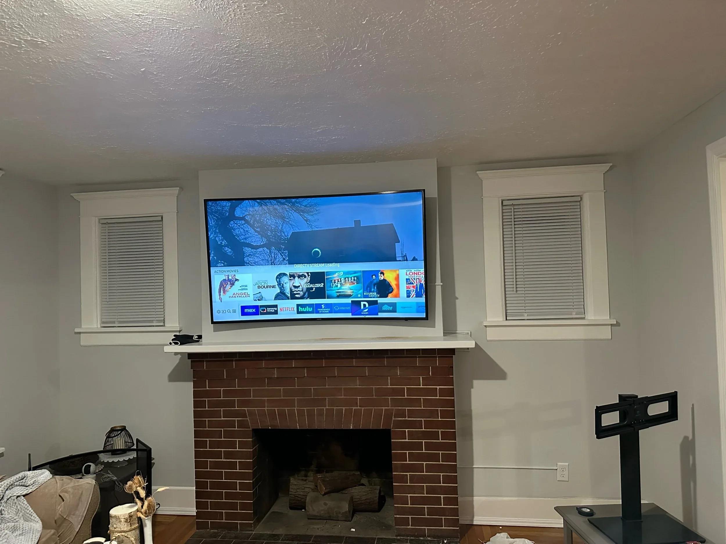 Flat-screen tv mounted above brick fireplace with white mantel and two windows with blinds