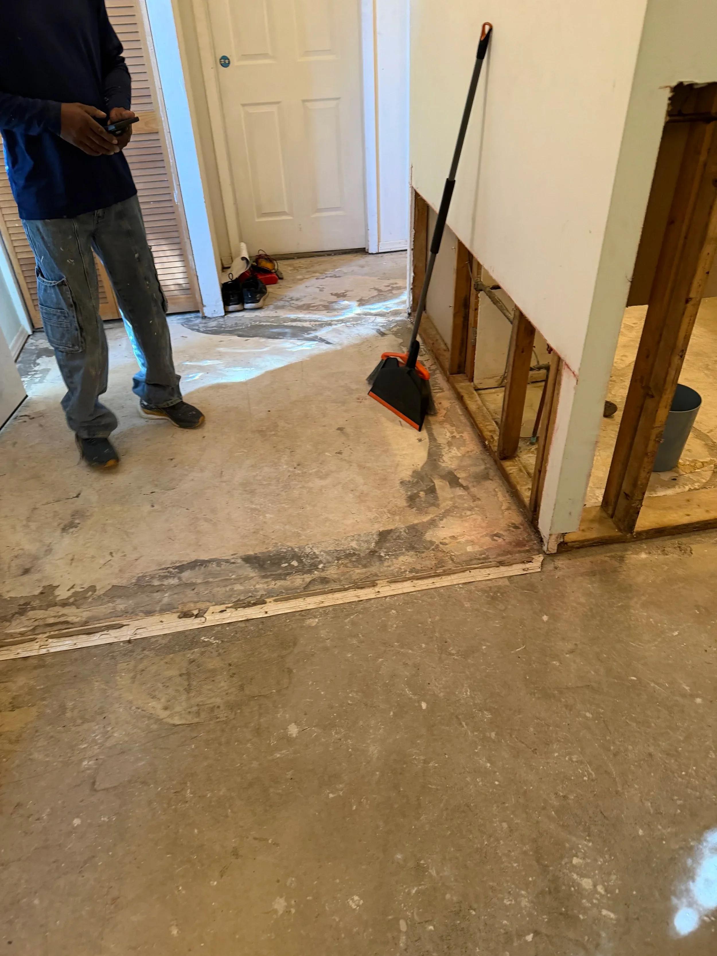 Worker standing next to broom and dustpan in gutted hallway with drywall removed from lower walls during renovation