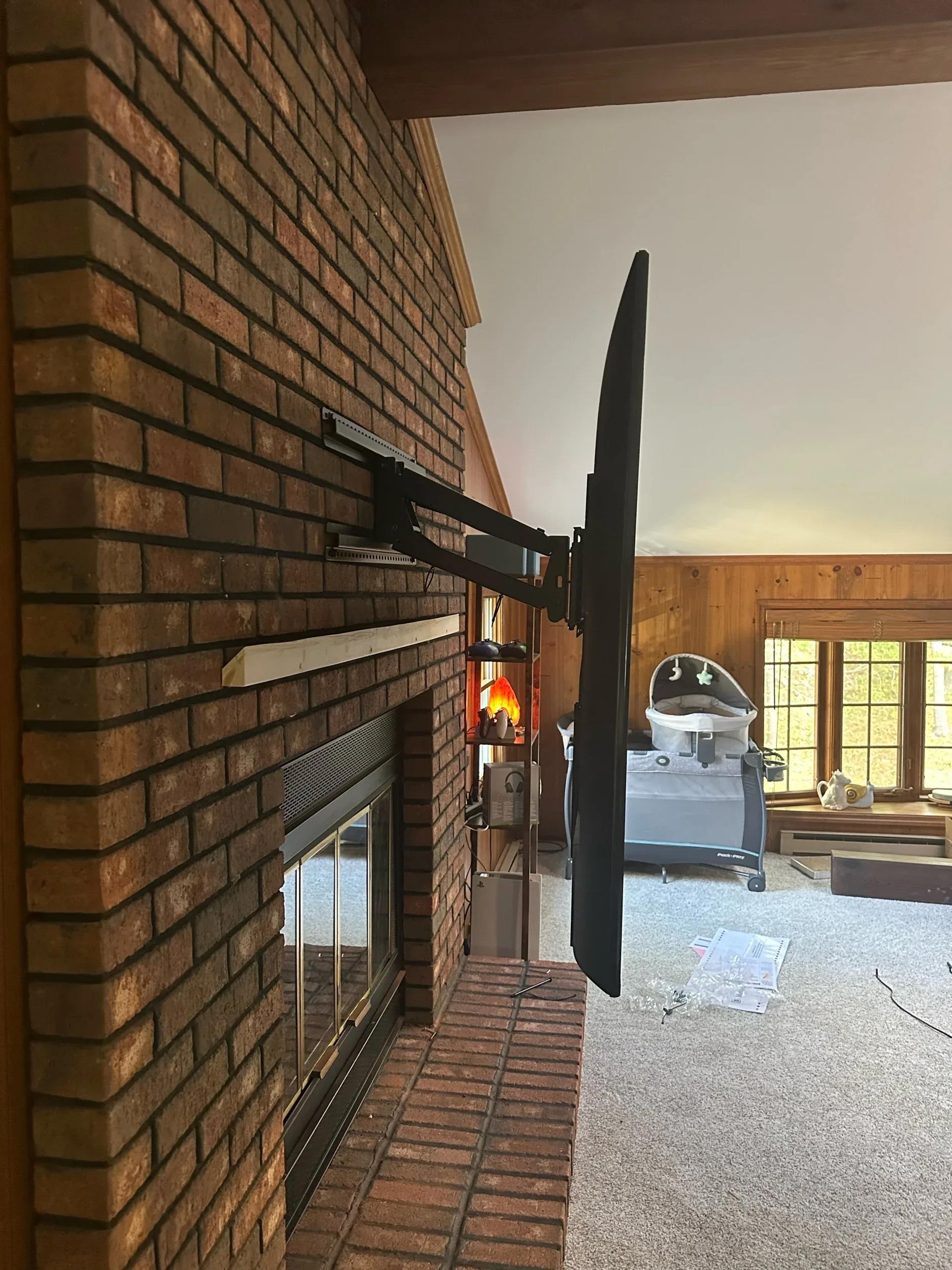 Flat-screen TV mounted on an extended swing-arm bracket above a brick fireplace in a wood-paneled room with nursery playpen