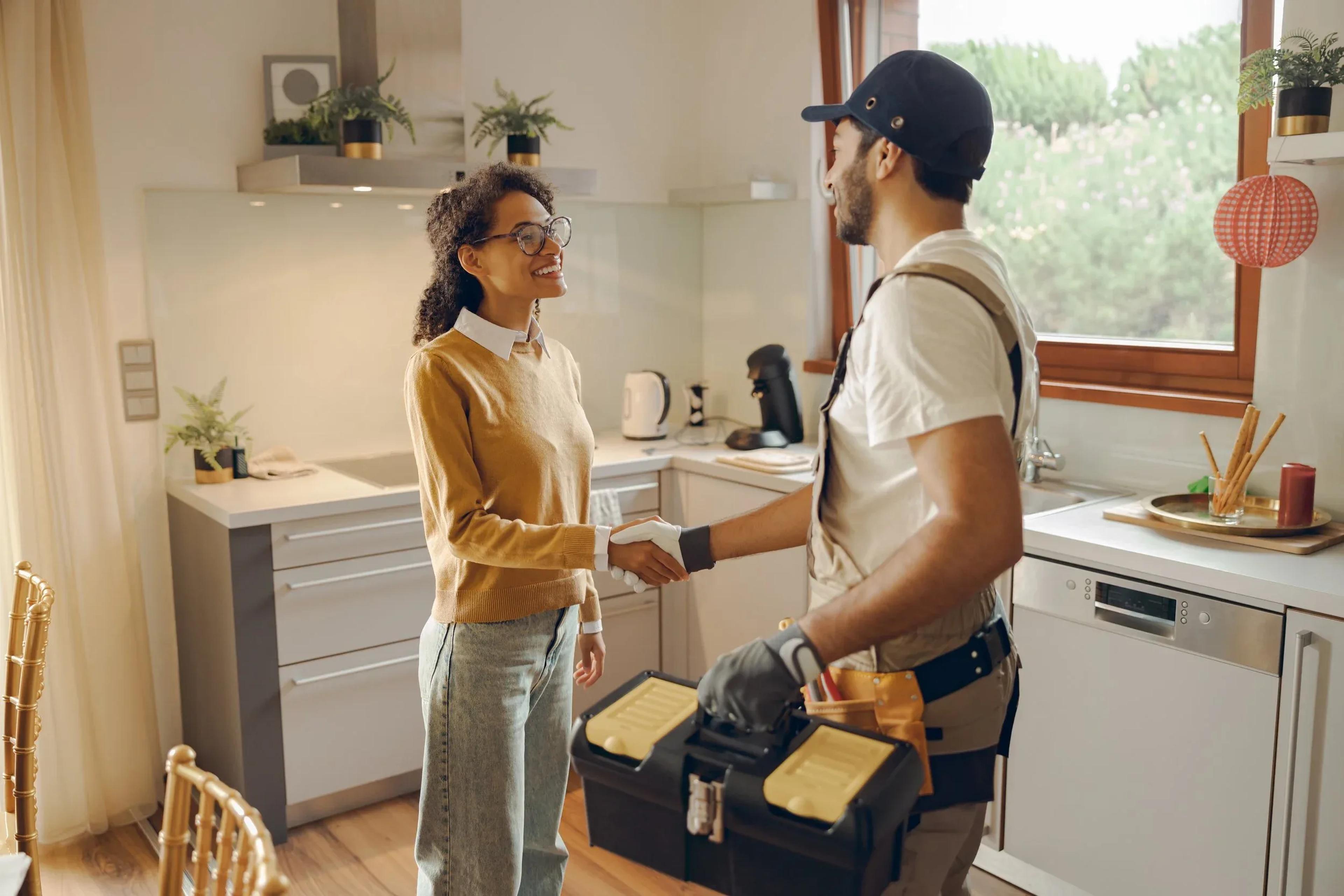 Electrician with tool bag shaking hands with homeowner in modern kitchen near dishwasher and window