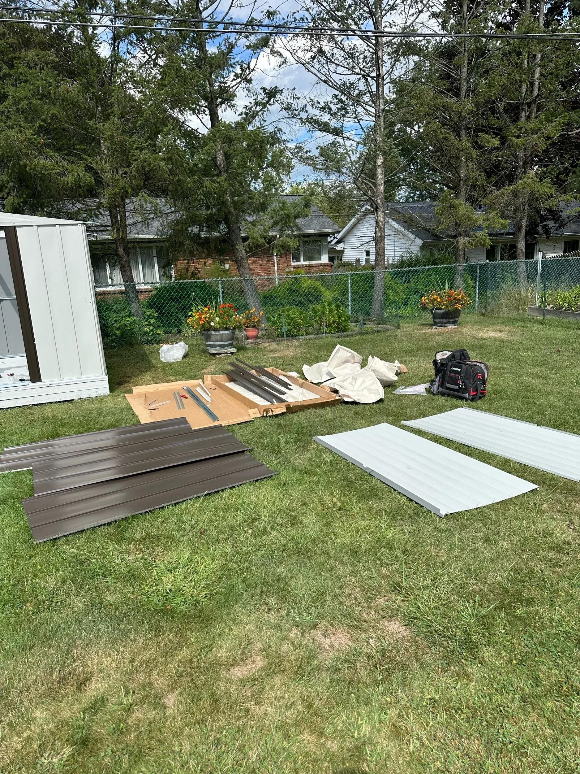 Metal shed panels, framing pieces, and tools laid out on grass for assembly near a partially built shed
