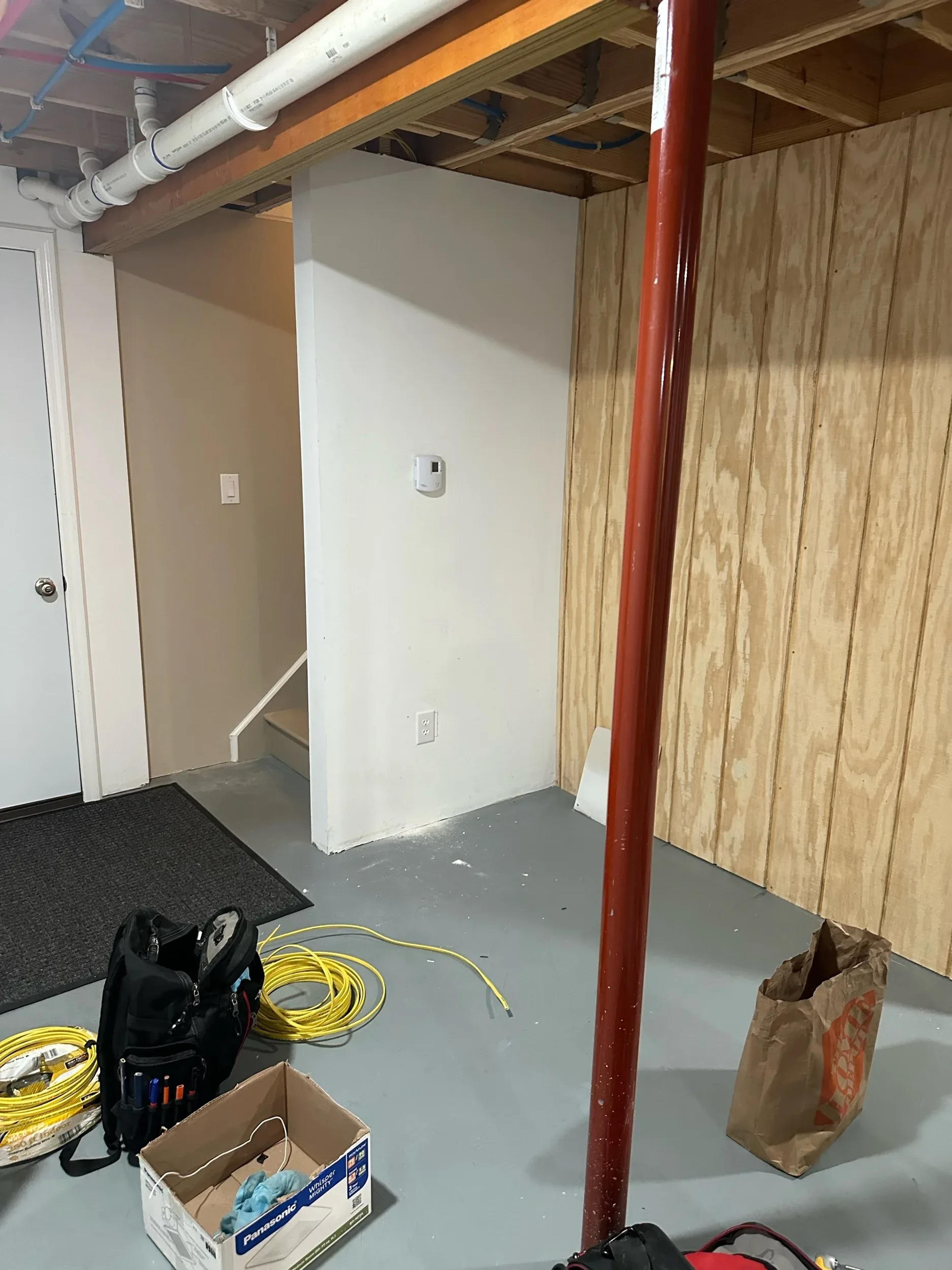 Unfinished basement corner with exposed ceiling joists, smoke detector, electrical outlet, tools, and coiled yellow wire