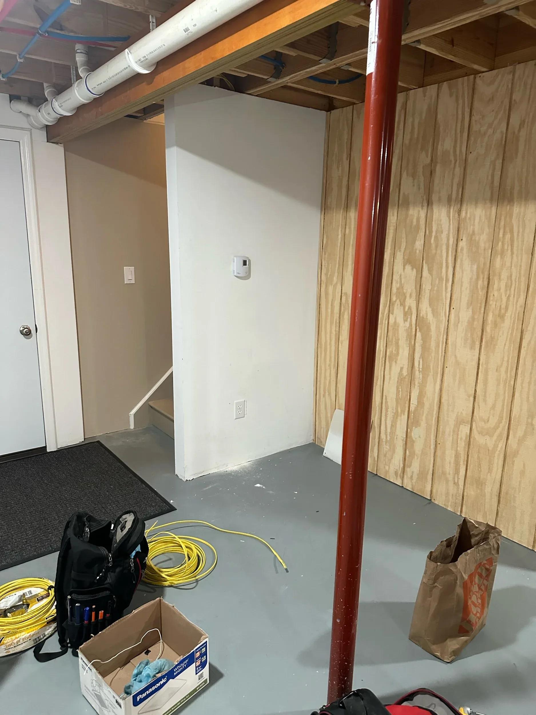 Unfinished basement corner with exposed ceiling joists, smoke detector, electrical outlet, tools, and coiled yellow wire