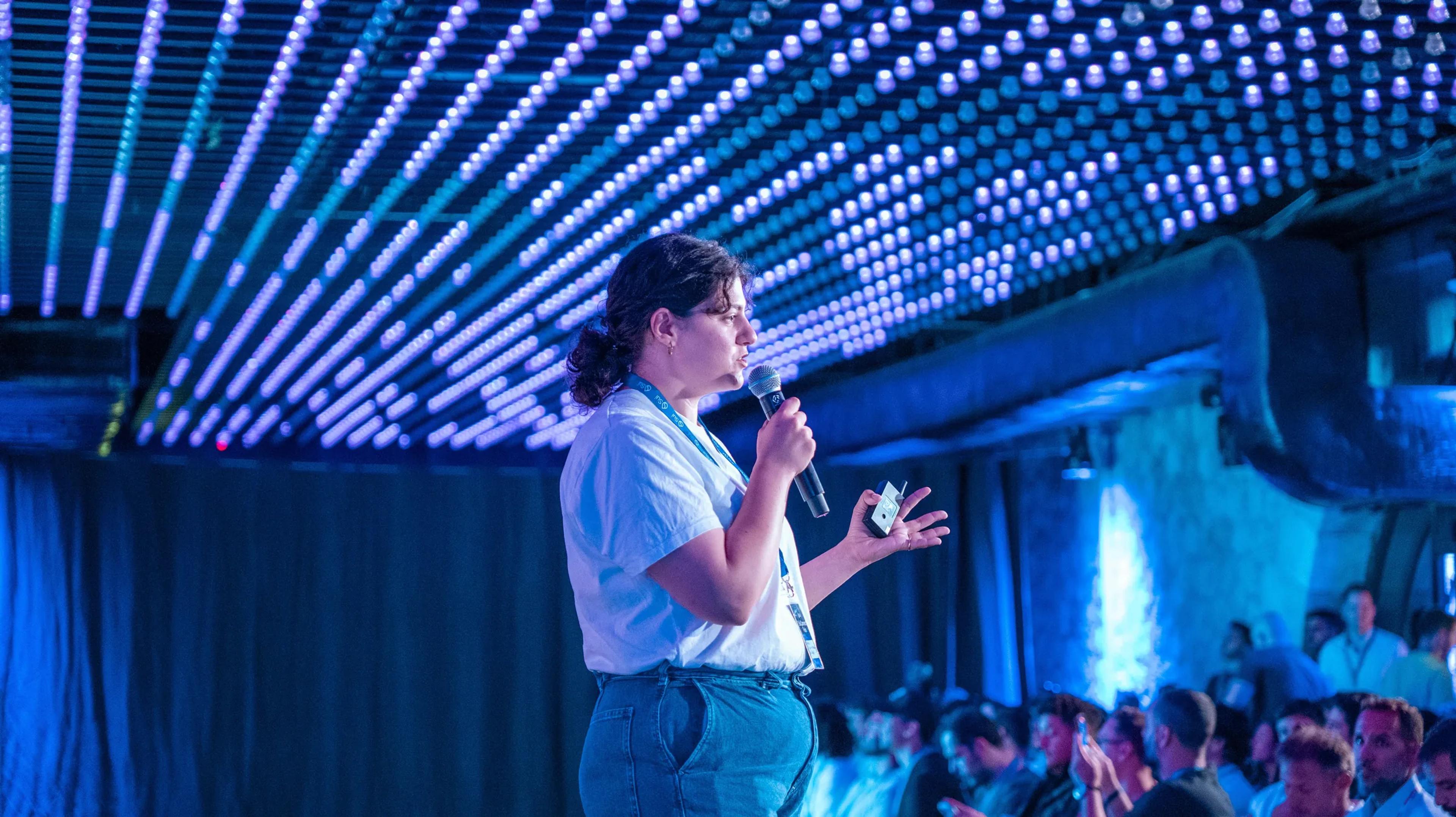 A speaker in front of a full room under blue stage lighting