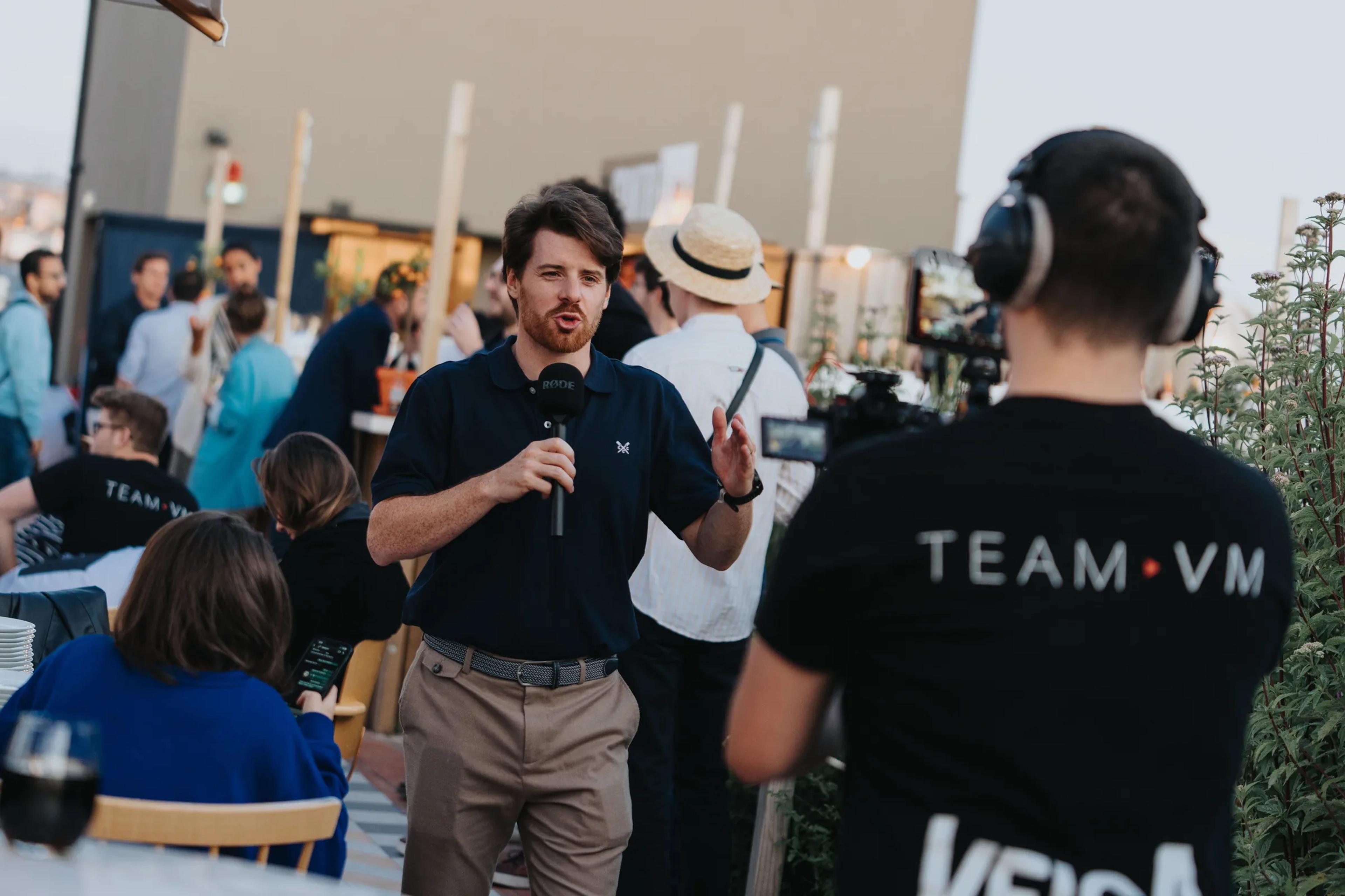 A host speaking into a microphone while a camera operator films