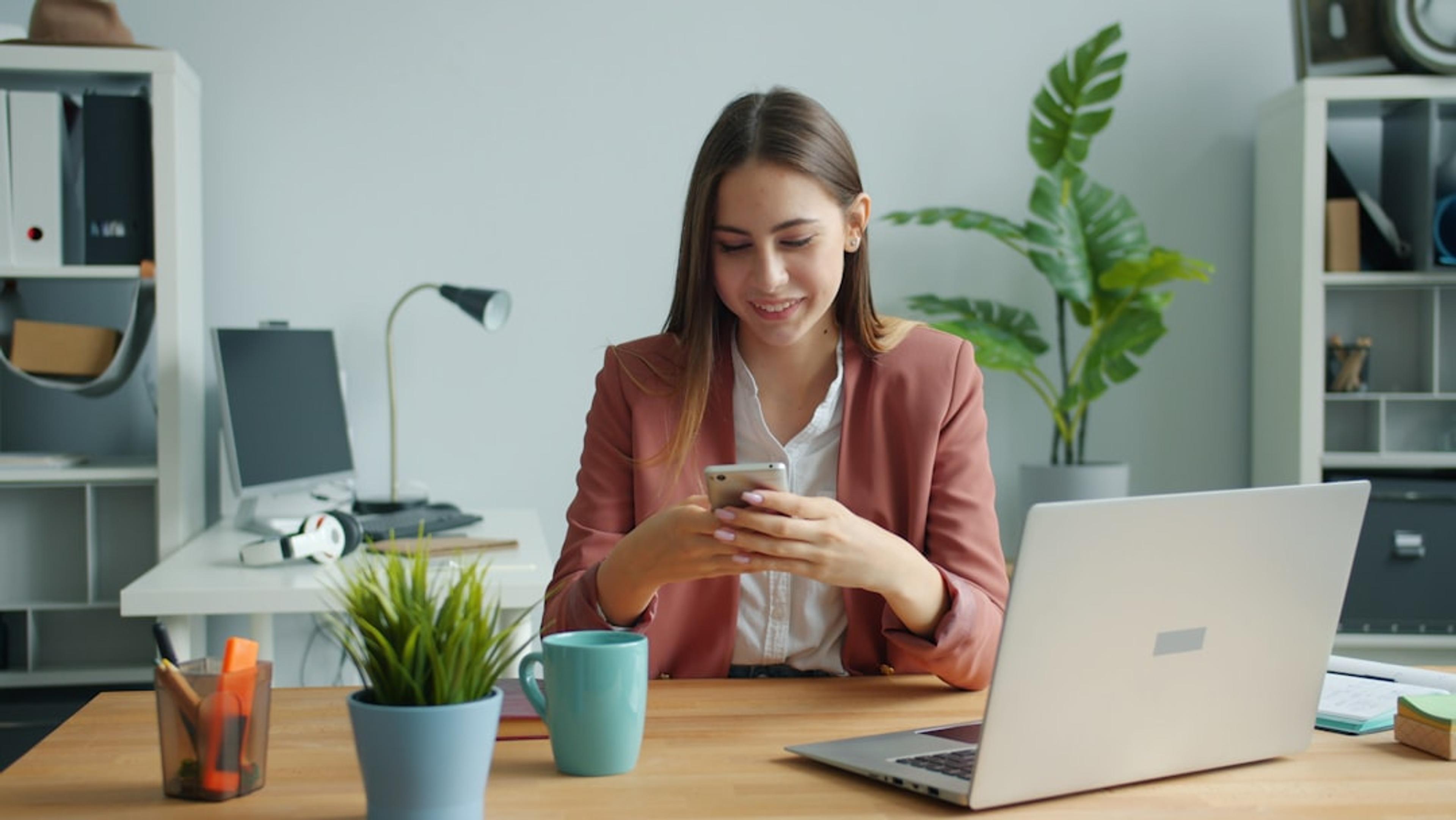 Beautiful lady is using smartphone touching screen smiling sitting at desk in office room alone enjoying gadget. People, workplace and devices concept.