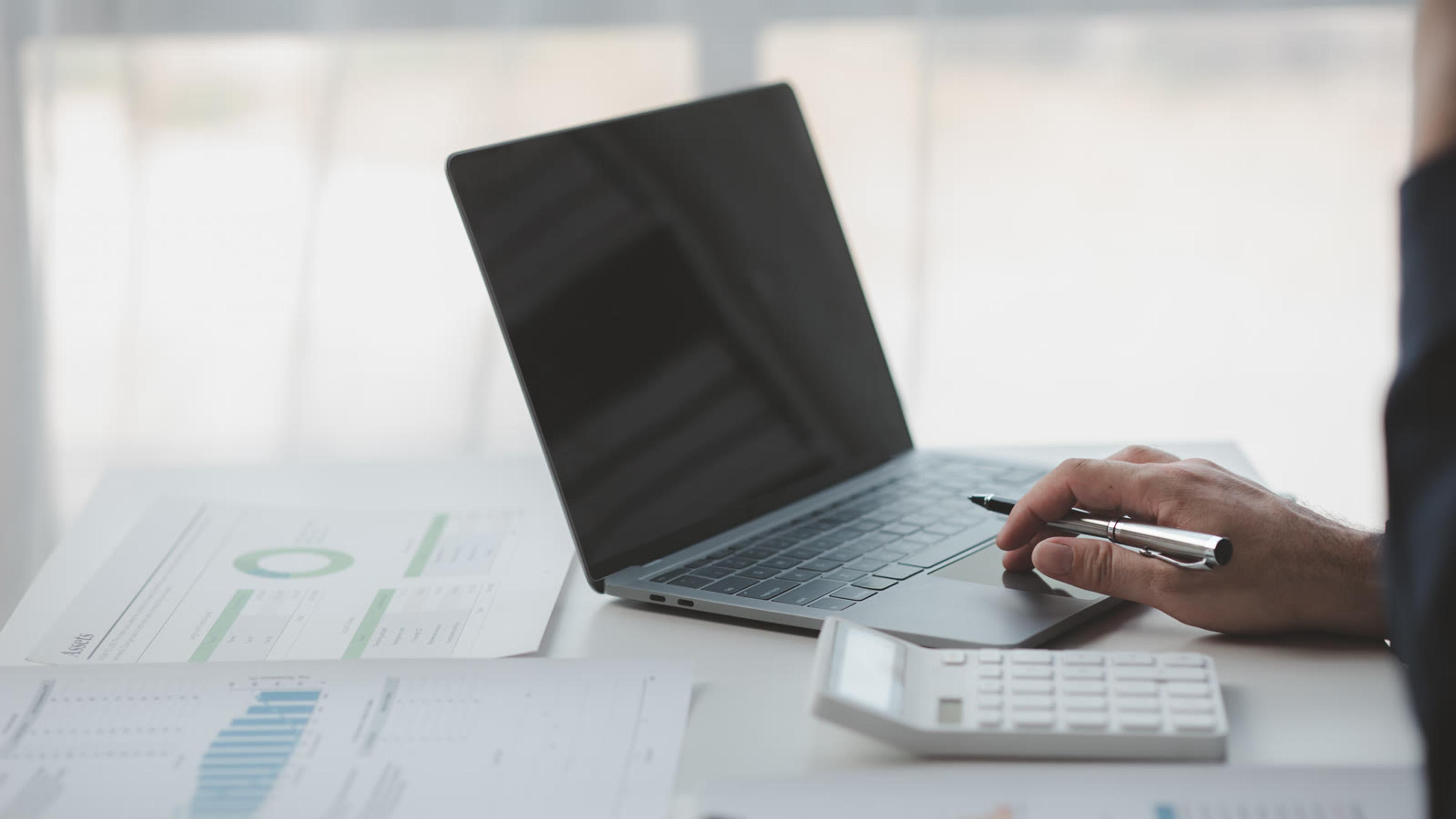 Hand using a laptop beside a calculator and printed cost charts while working out voice margins