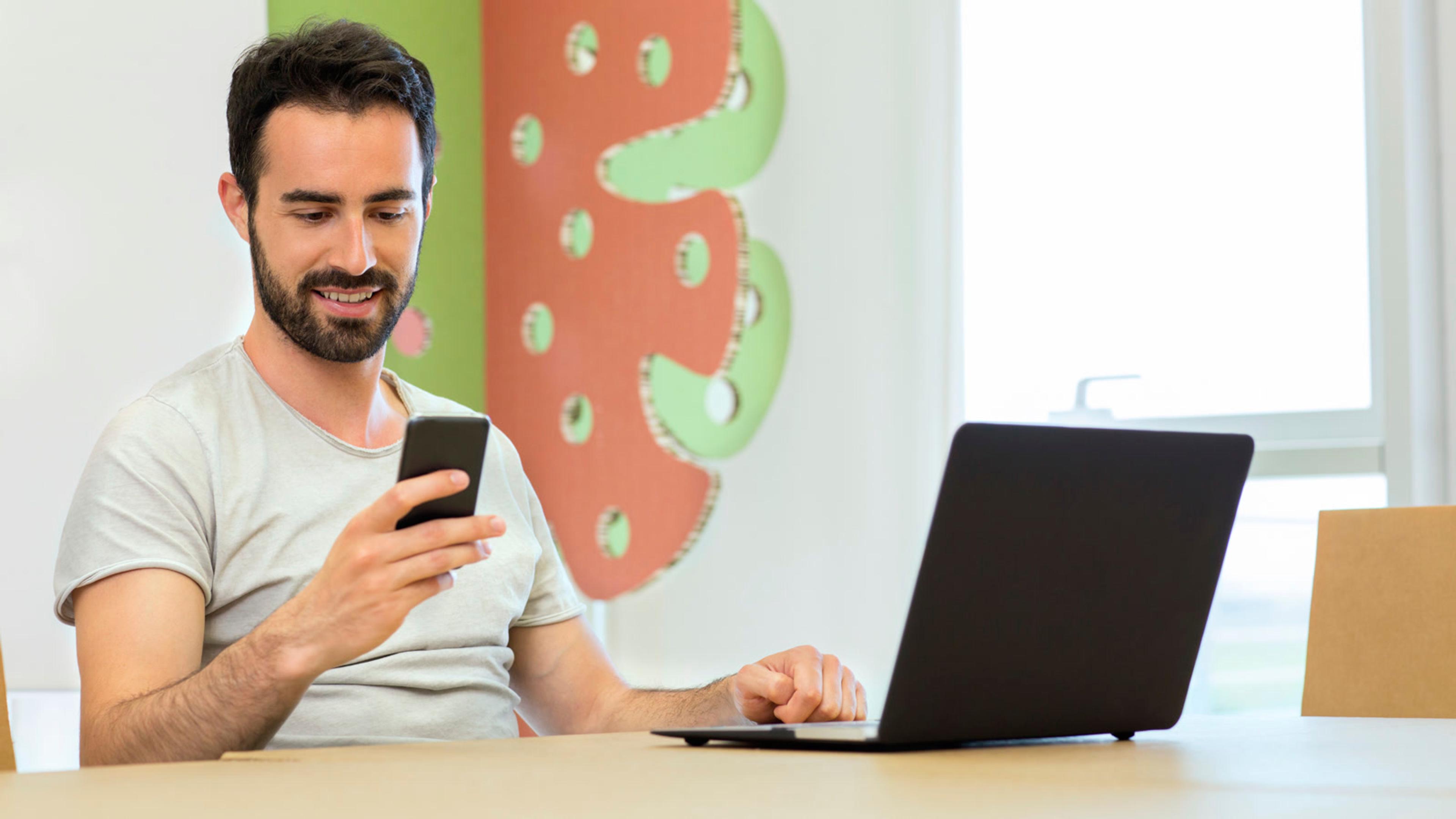 Man with phone and laptop at a desk — BYOD softphone on a personal device