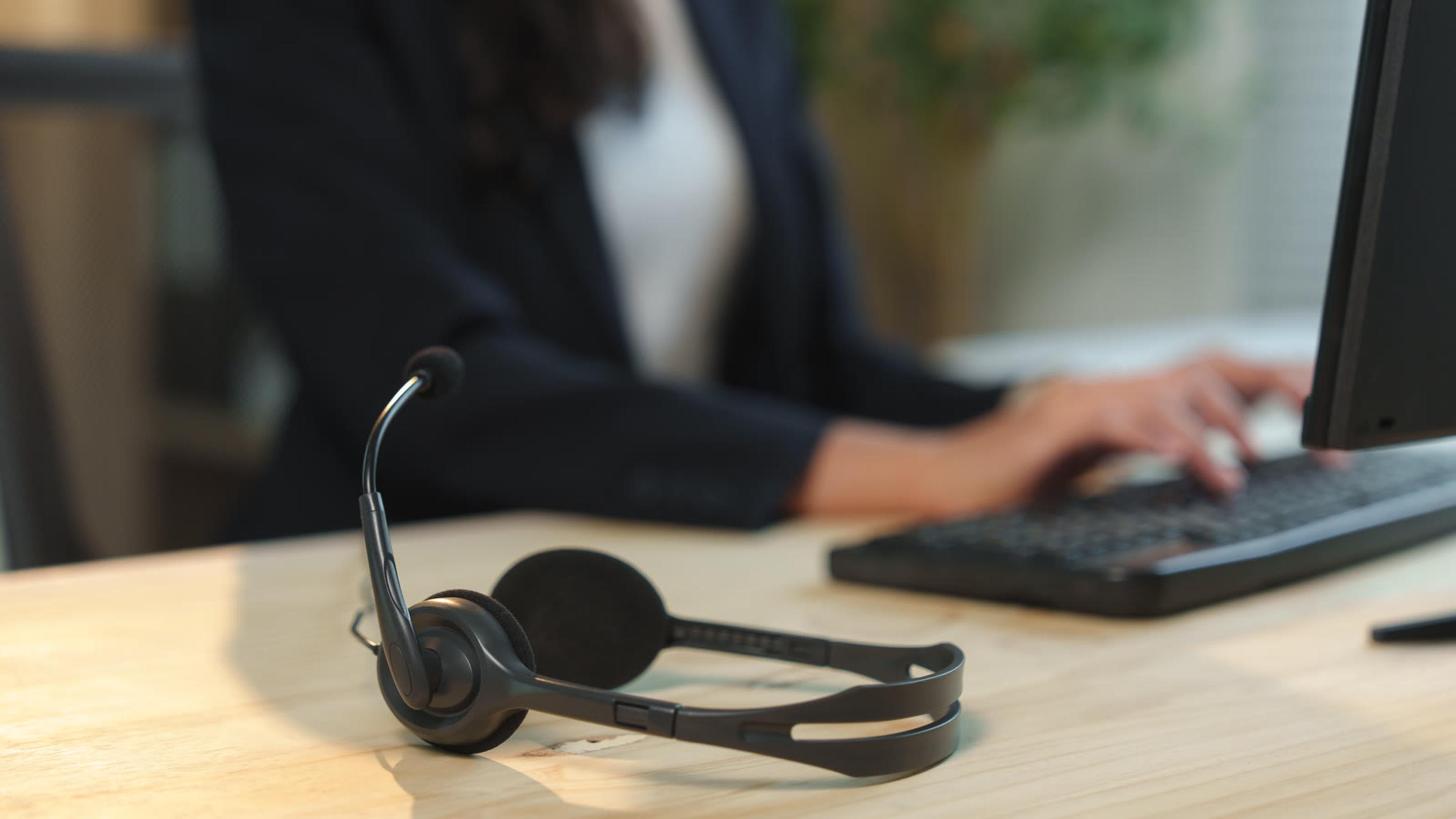 Headset on an office desk while a colleague types, ready to take calls through a desktop softphone