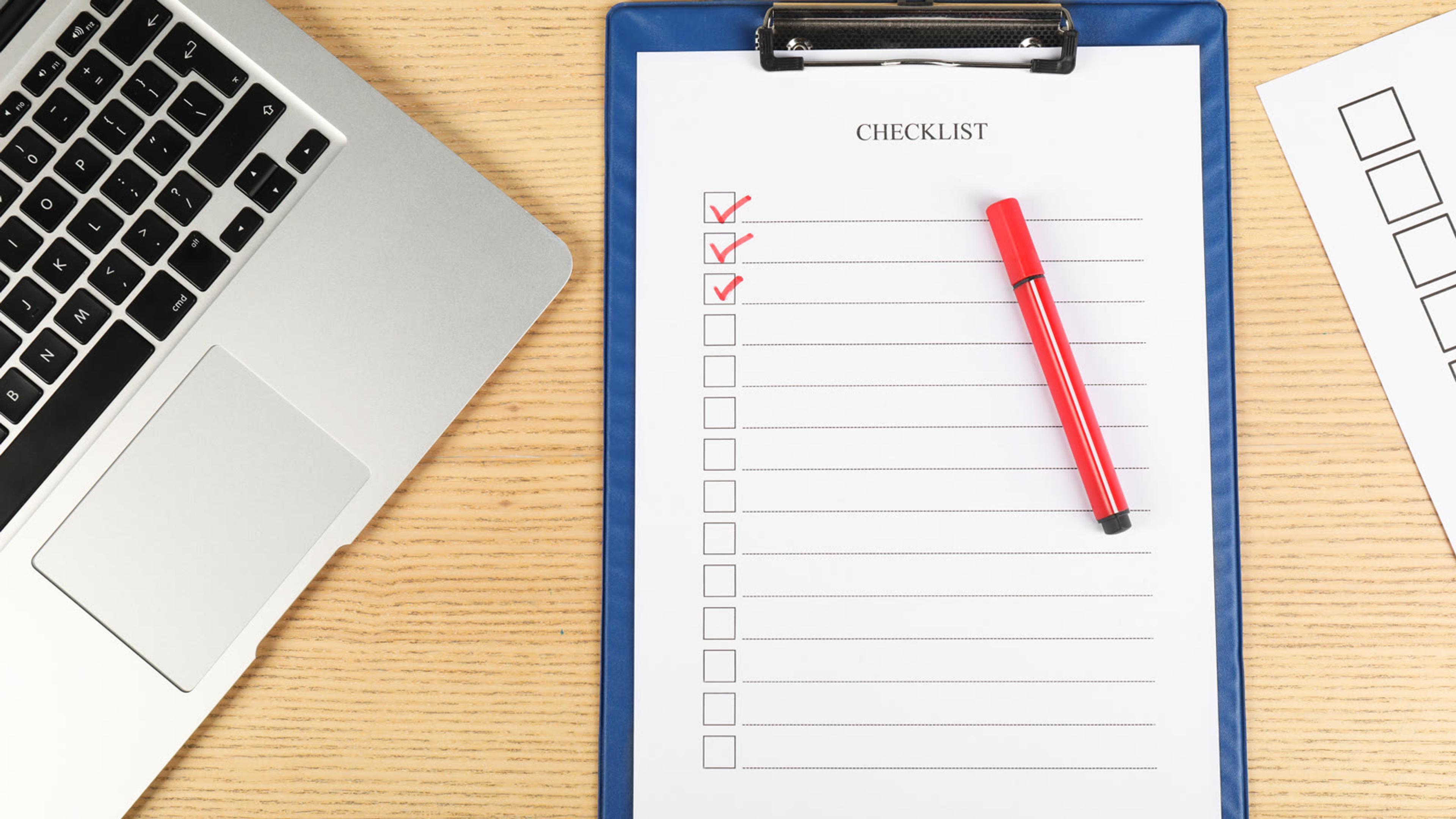 Clipboard with a partly ticked checklist and a red pen on a desk beside a laptop