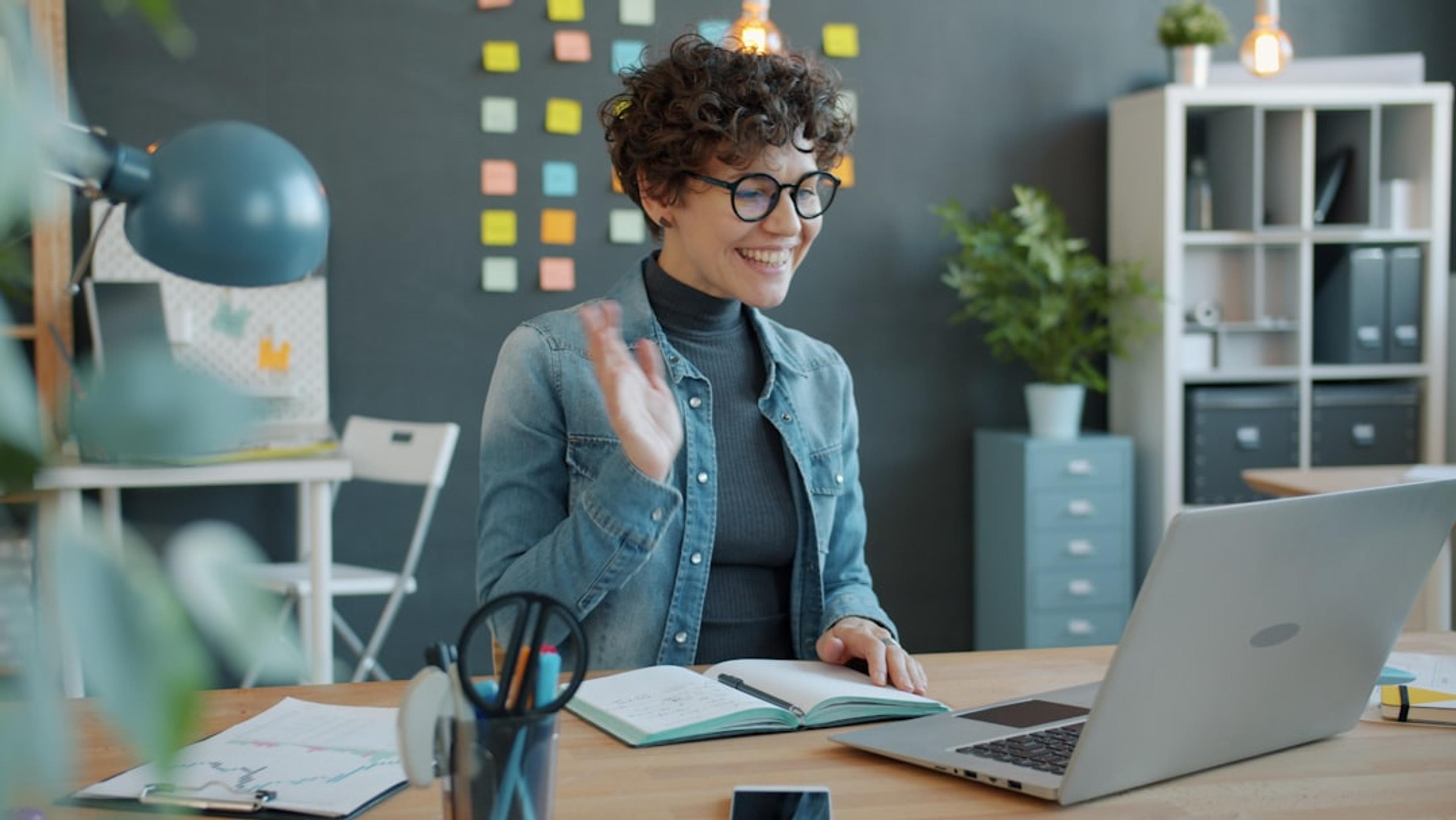 Remote worker waving during a video call at a home office desk