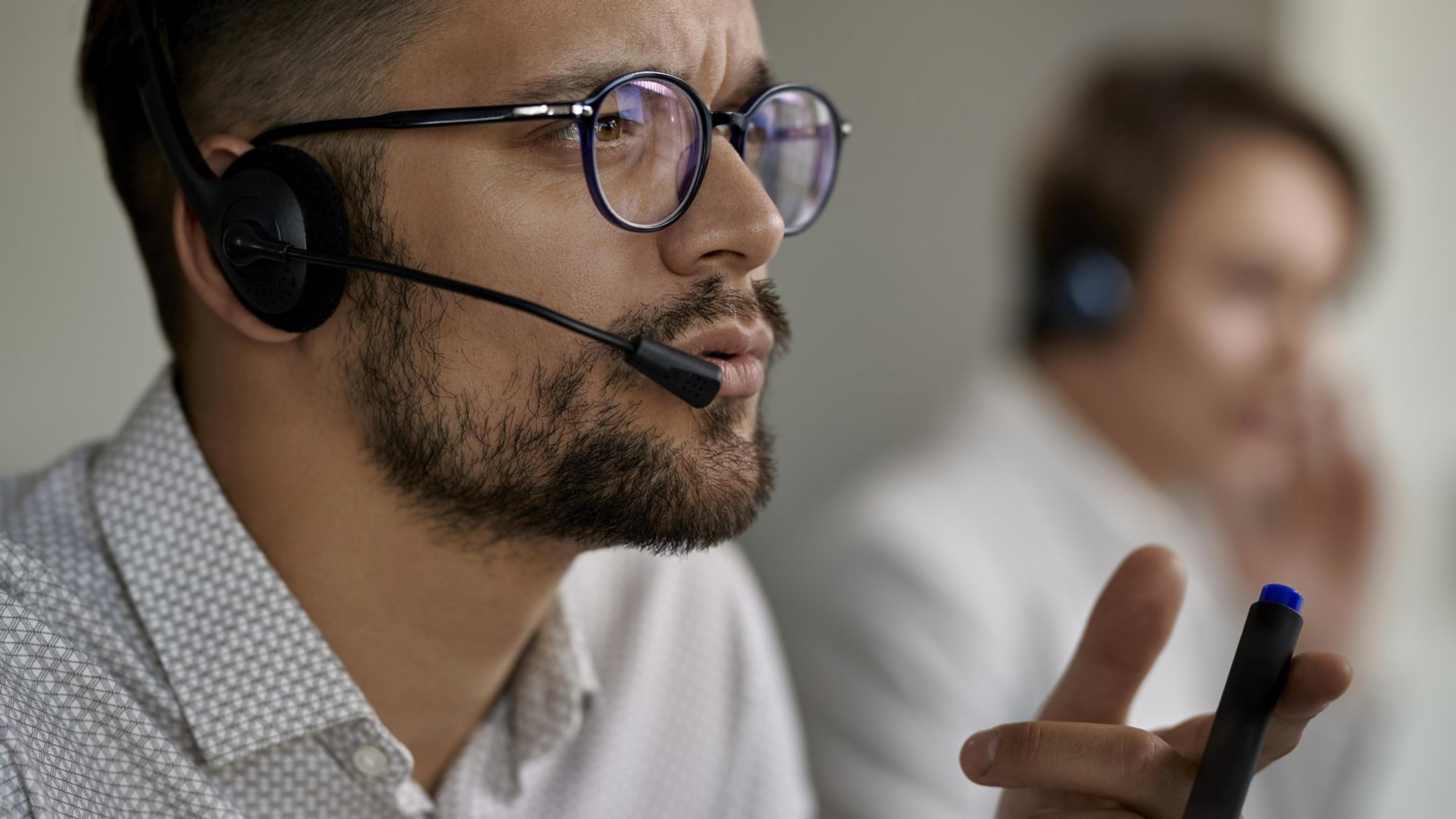Customer service agent wearing a headset and glasses concentrating on a call, with a colleague on a headset behind him