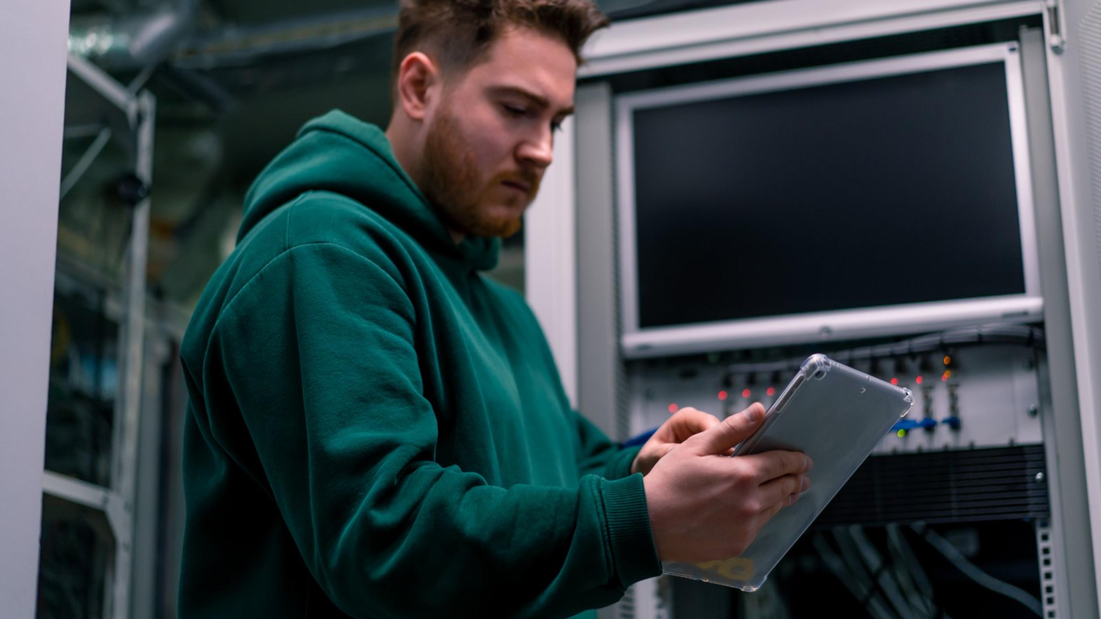 IT administrator in a green hoodie checking a tablet while standing at a server rack