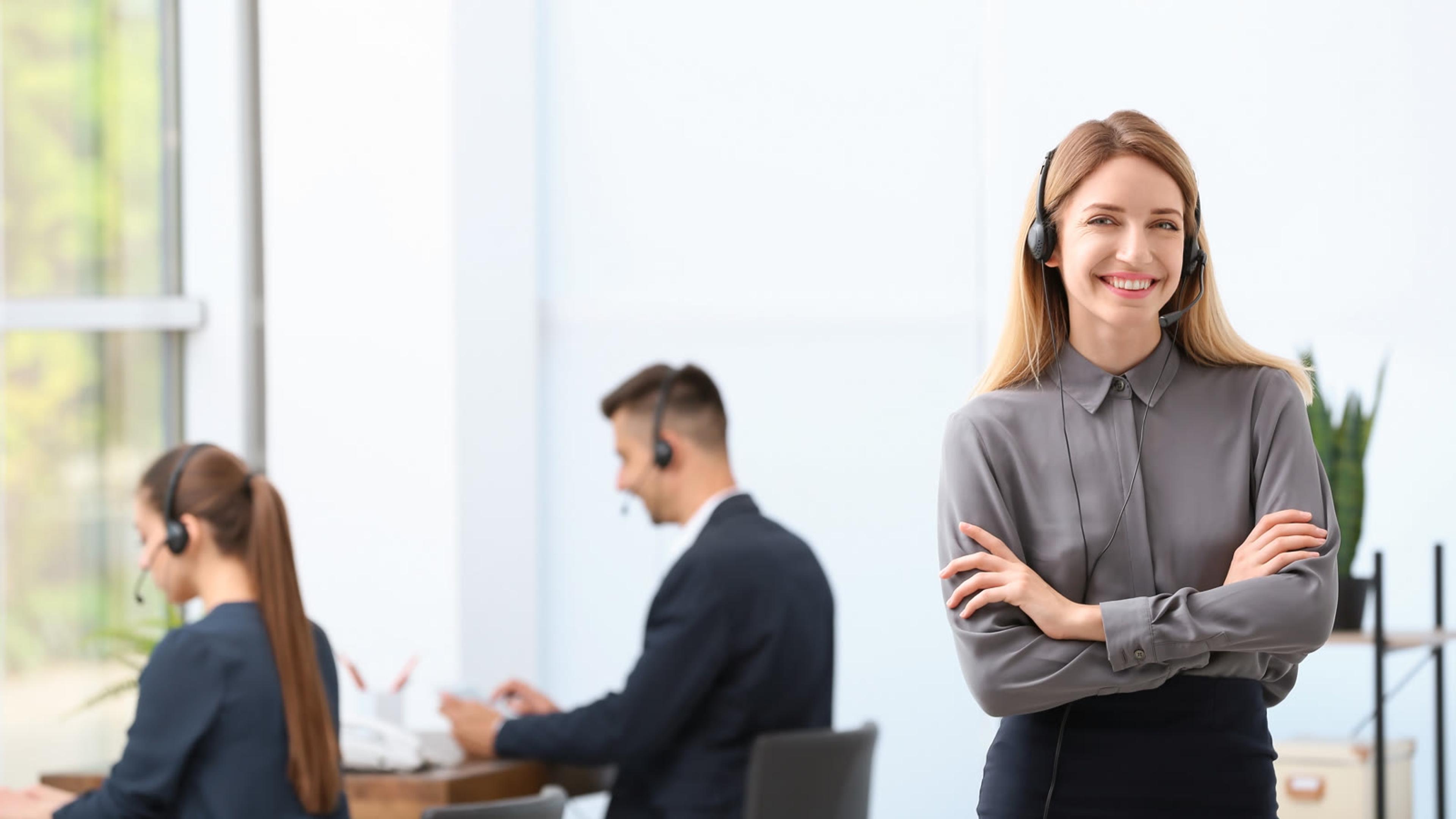 Team lead wearing a headset stands in the office while two colleagues in headsets work behind her