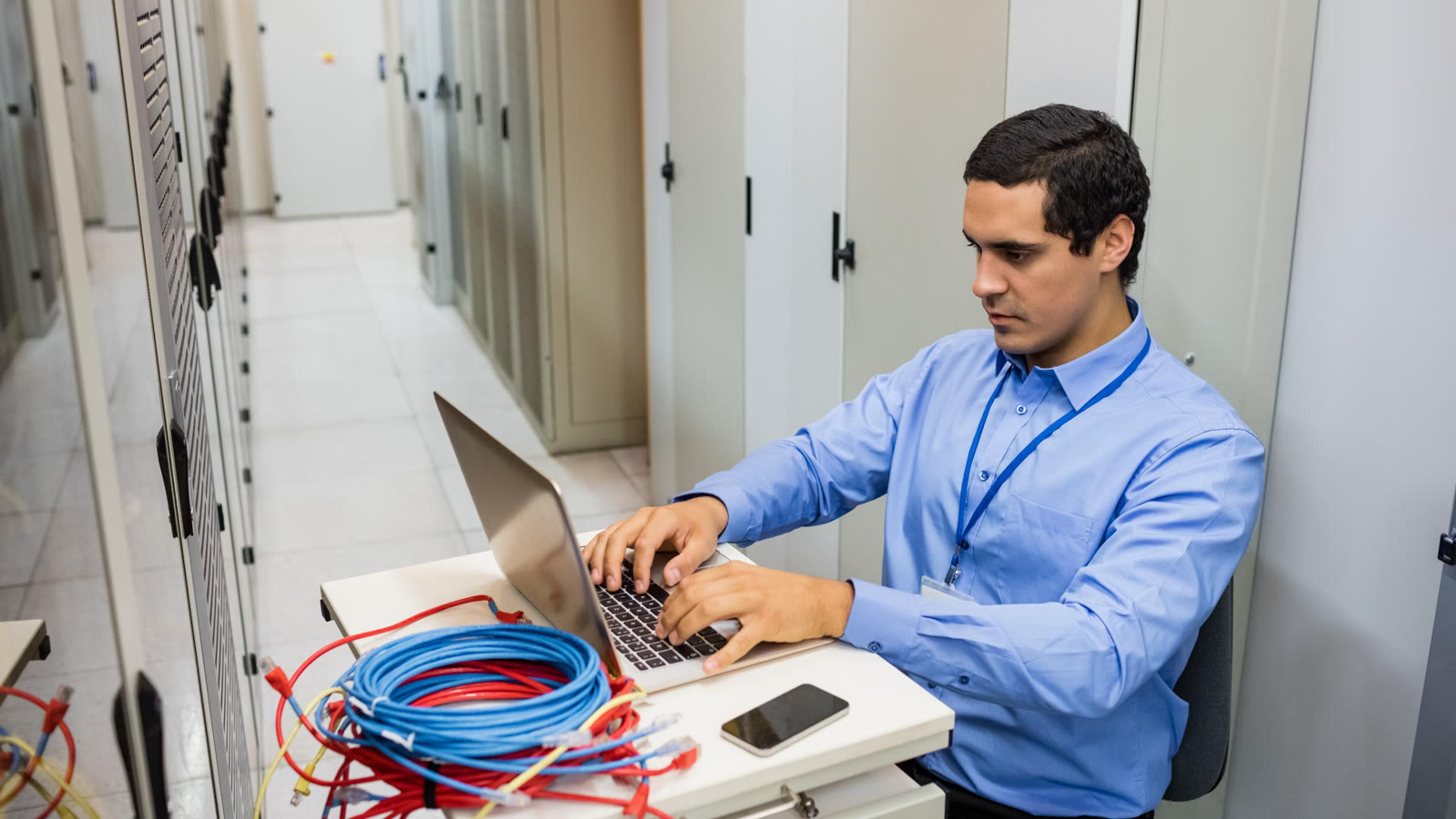 IT technician working on a laptop in a server room while preparing a softphone rollout